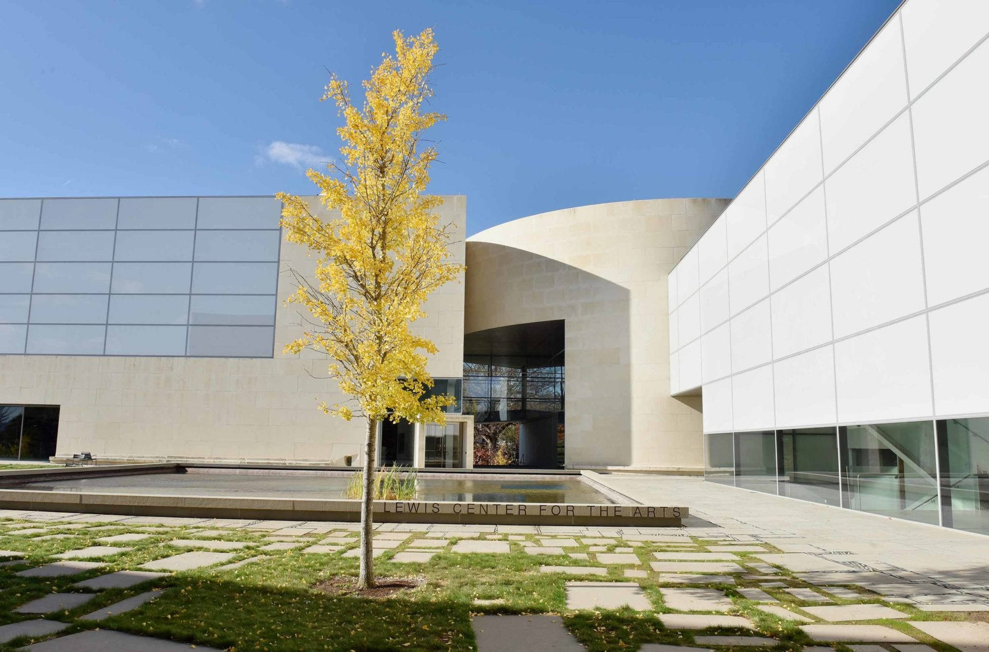 Building 桌布 壁紙 — 一座現代建築，擁有彎曲的米色牆壁和玻璃面板，在晴朗的藍天下，修剪整齊的庭院中有一棵充滿活力的黃色樹木。