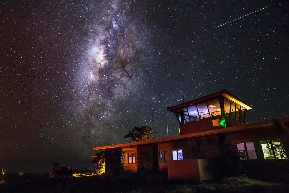 Night Time 桌布 壁紙 — 一座現代建築在壯麗的星空下溫暖地發光，銀河拱橋般橫跨上空。