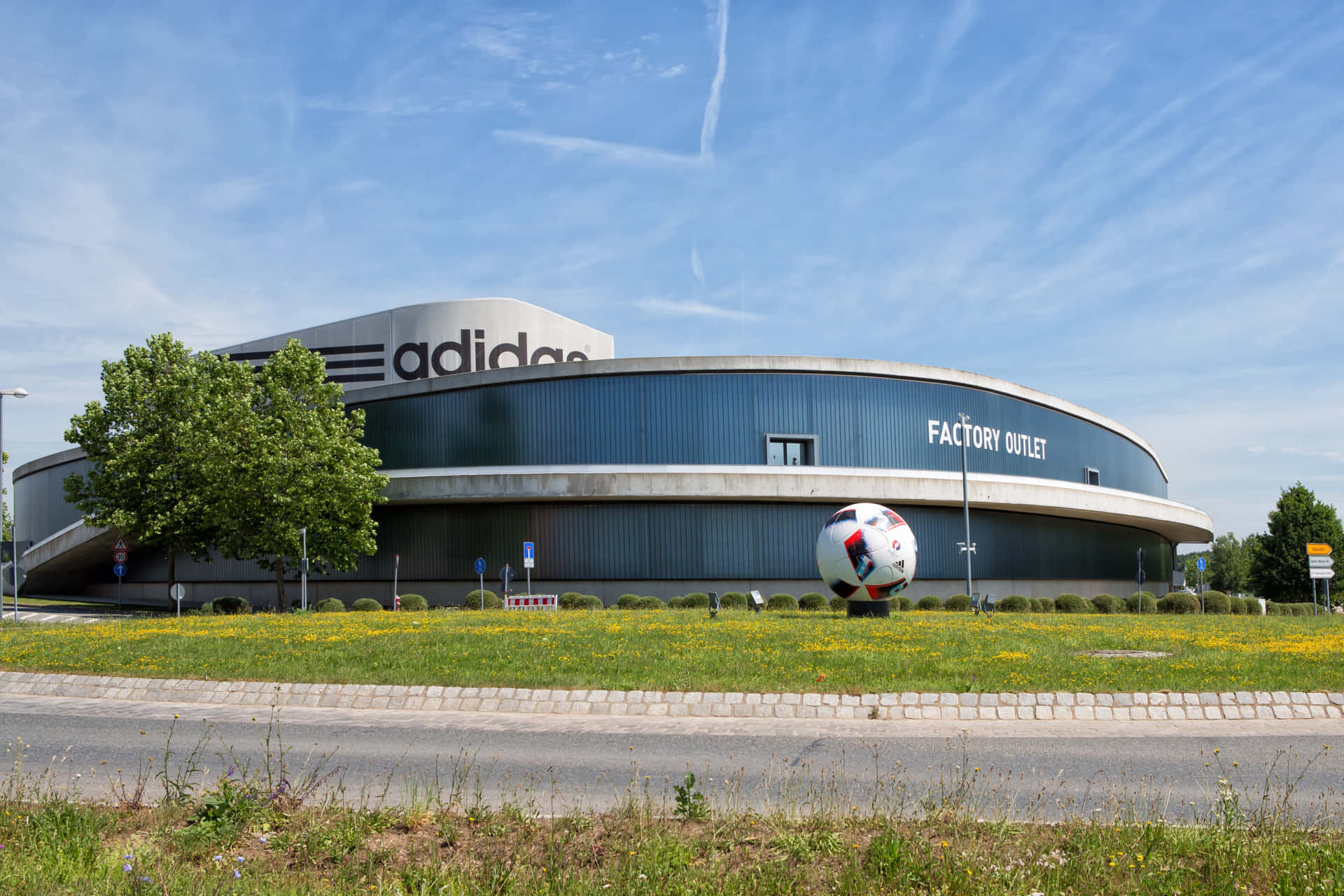 愛迪達 桌布 壁紙 — 一座現代弧形 adidas 工廠直銷店建築，在明亮的藍天下，前面有一個巨大的足球雕塑。