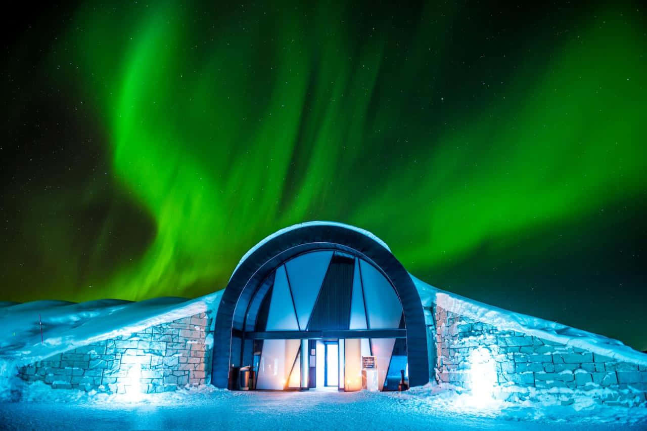 Ice Hotel 桌布 壁紙 — 一座現代拱形建築物坐落在雪地中，上方有 vibrant 綠色極光在繁星點點的夜空中舞動。