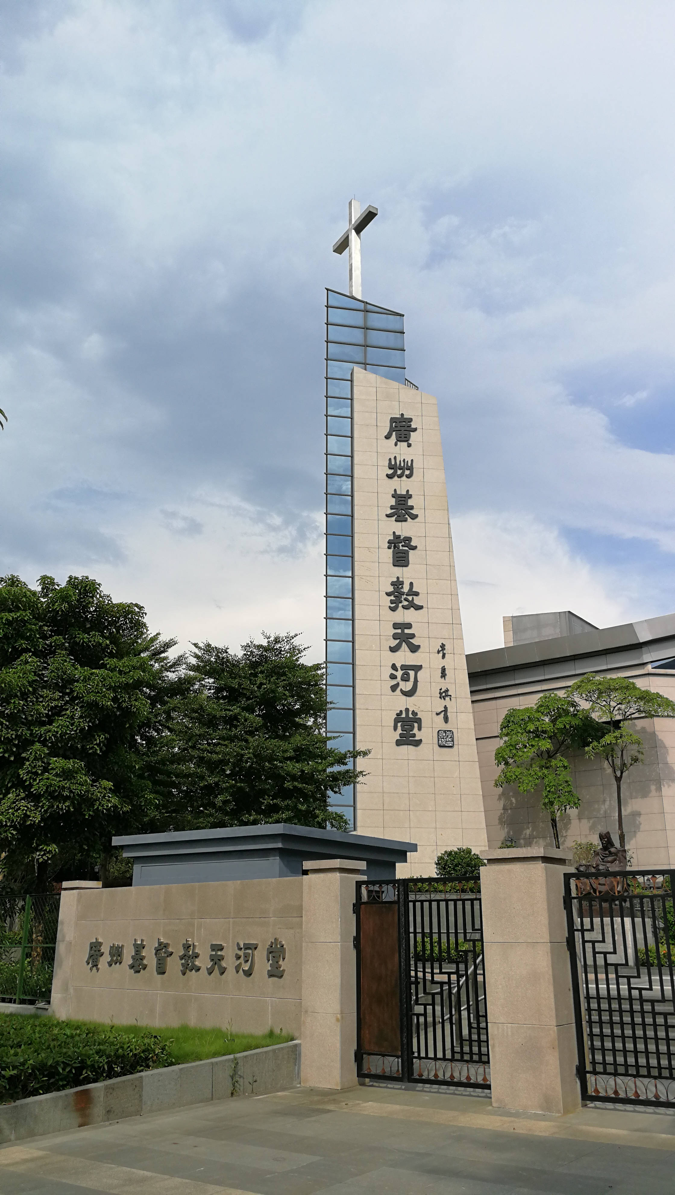 Guangzhou 桌布 壁紙 — 一座現代教堂，擁有高聳的玻璃和石頭塔樓，頂端有十字架，矗立在多雲的天空下，茂密的綠樹環繞著有門的入口。