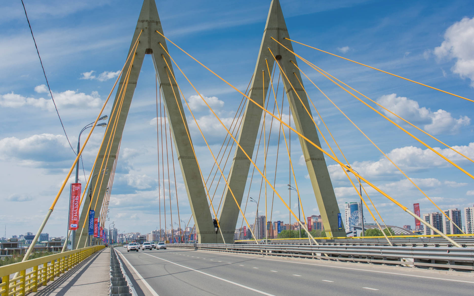 Kazan 桌布 壁紙 — 一座現代斜張橋，黃色纜索橫跨城市景觀，天空湛藍，白雲朵朵。