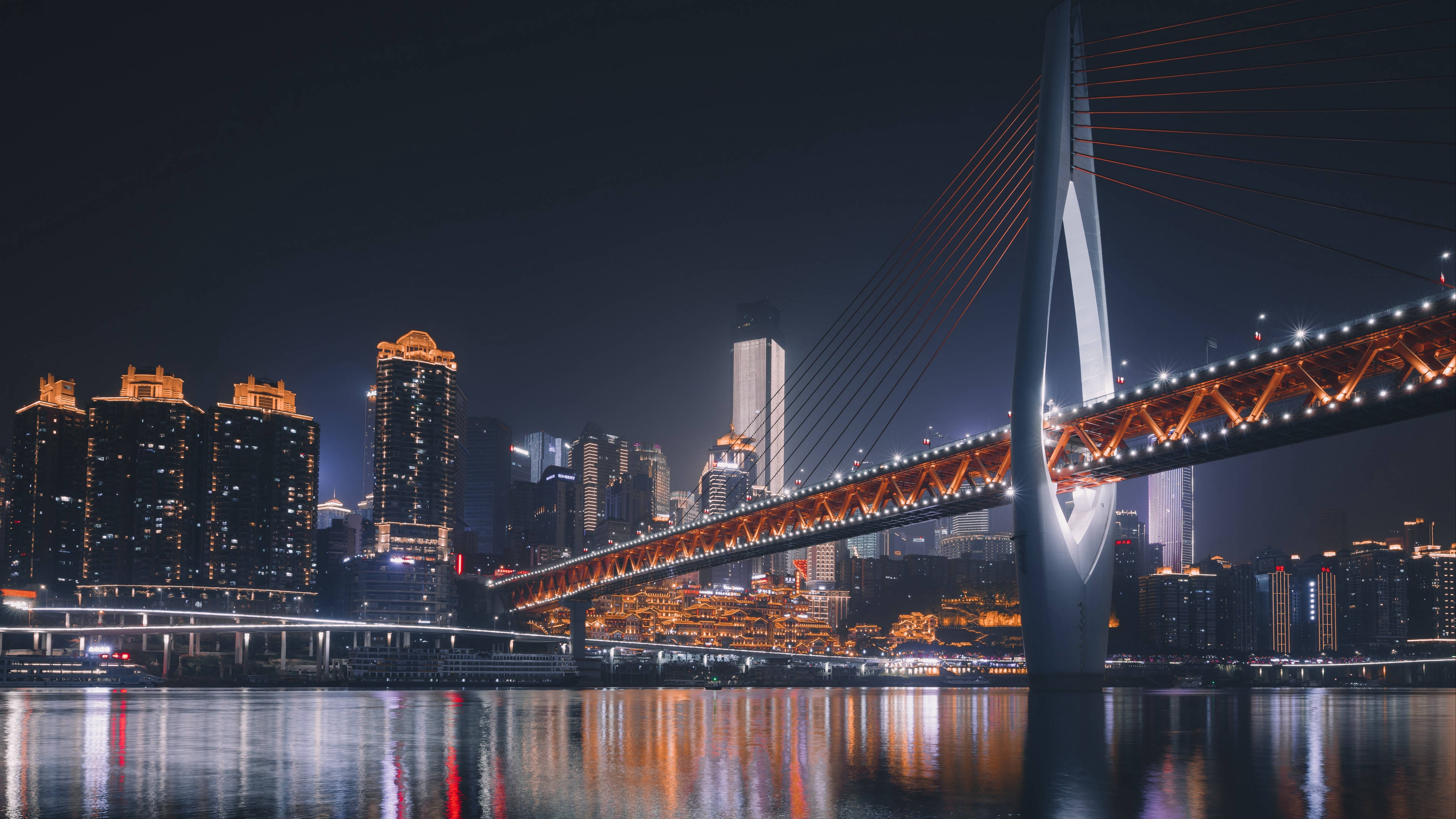 Chongqing 桌布 壁紙 — 一座現代斜張橋在反射的河流上閃爍著溫暖燈光的迷人夜景，被密集的城市天際線所環繞。