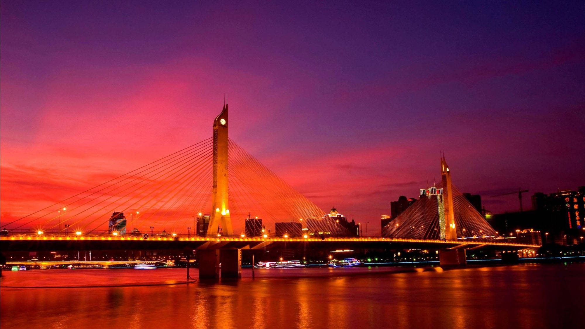 Guangzhou 桌布 壁紙 — 一座現代斜張橋在平靜的河面上，以溫暖的燈光映襯著火紅的日落天空。