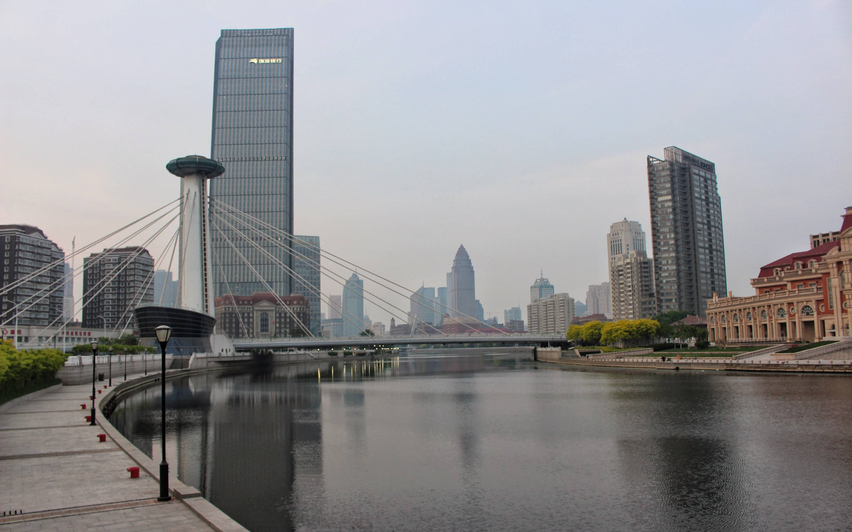 Tianjin 桌布 壁紙 — 一座現代斜張橋橫跨平靜的河流，摩天大樓和歷史建築沿著海濱排列，天空多雲。