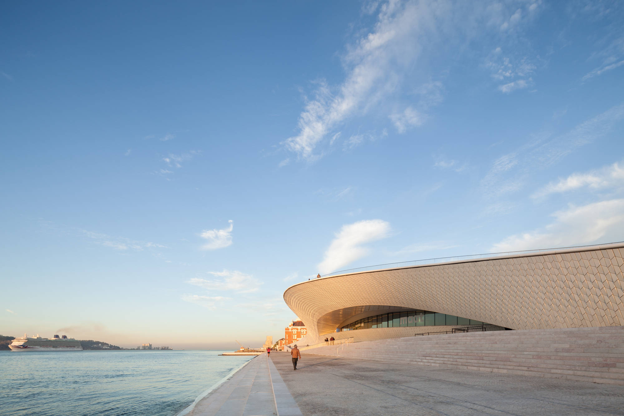Museum 桌布 壁紙 — 一座現代曲線建築，帶有紋理外牆，矗立在平靜的水邊，天空清澈湛藍，飄著縷縷白雲。
