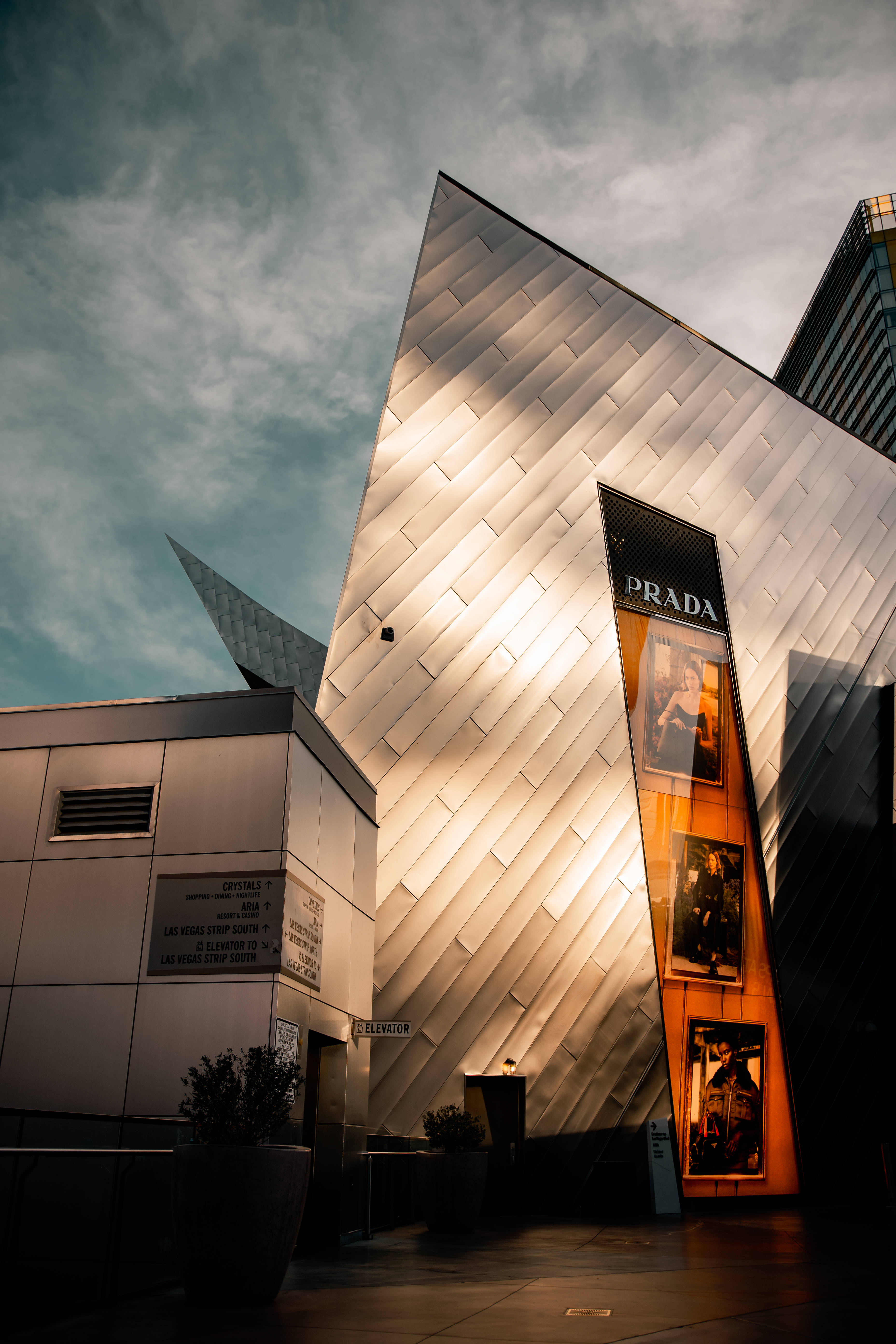Prada 桌布 壁紙 — 一座現代有稜角的Prada建築，帶有發光面板和溫暖的內部光芒，映襯著多雲的黃昏天空。