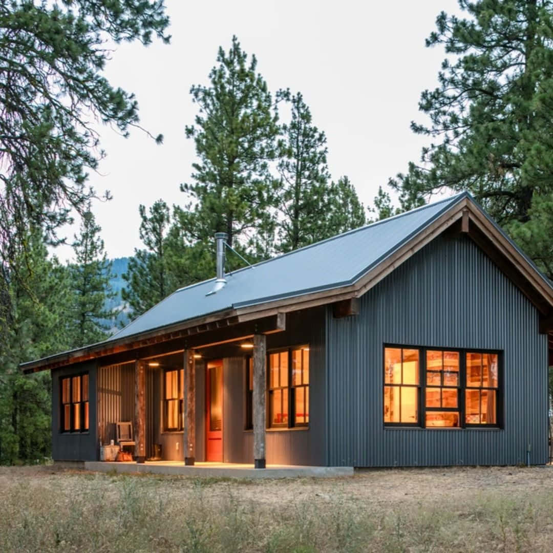 Cottage 桌布 壁紙 — 一座現代深灰色小屋坐落在松樹林中，黃昏時溫暖的室內燈光透過窗戶閃爍。