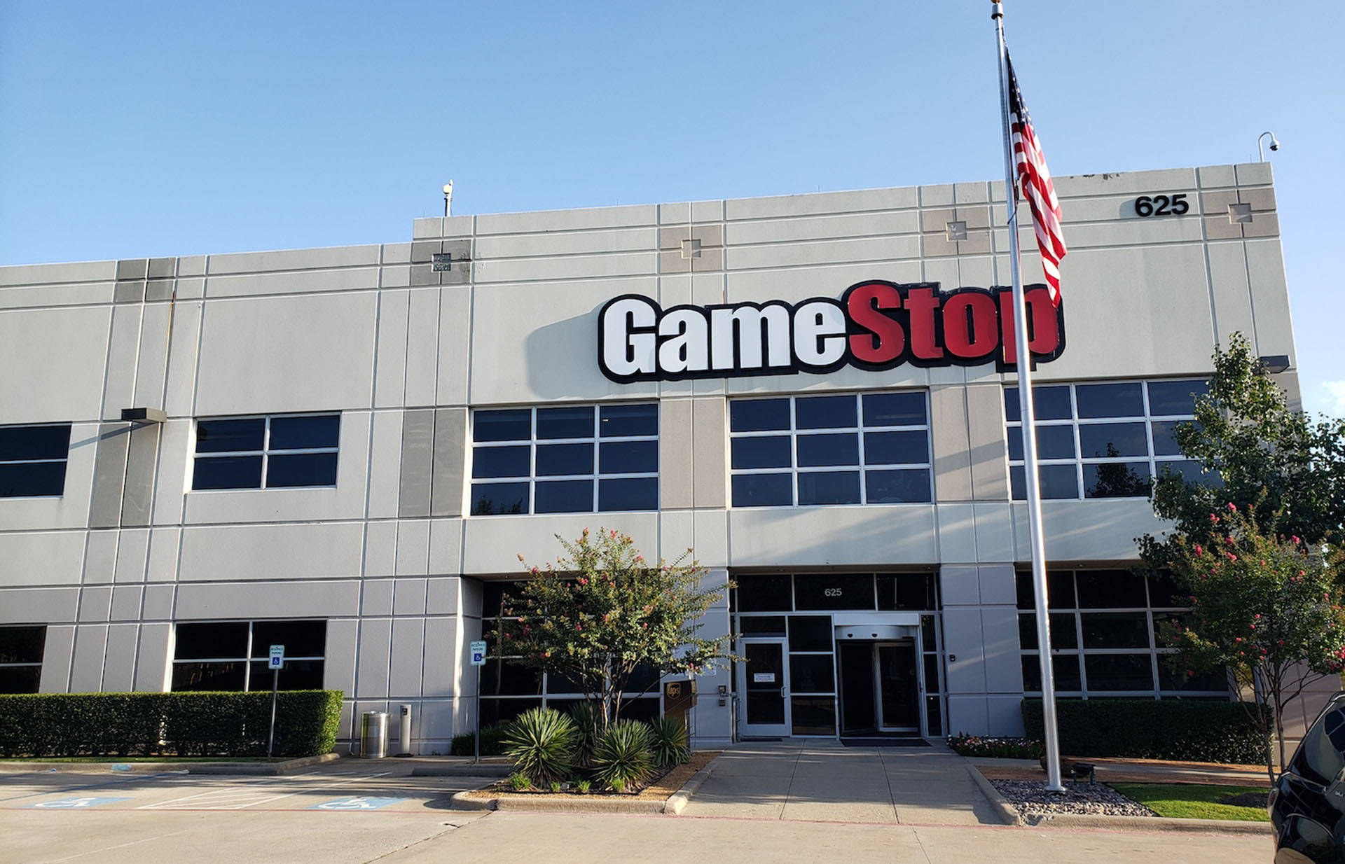 GameStop 桌布 壁紙 — 一座現代灰色GameStop商店建築，在晴朗藍天下，配有美國國旗和綠色園景。