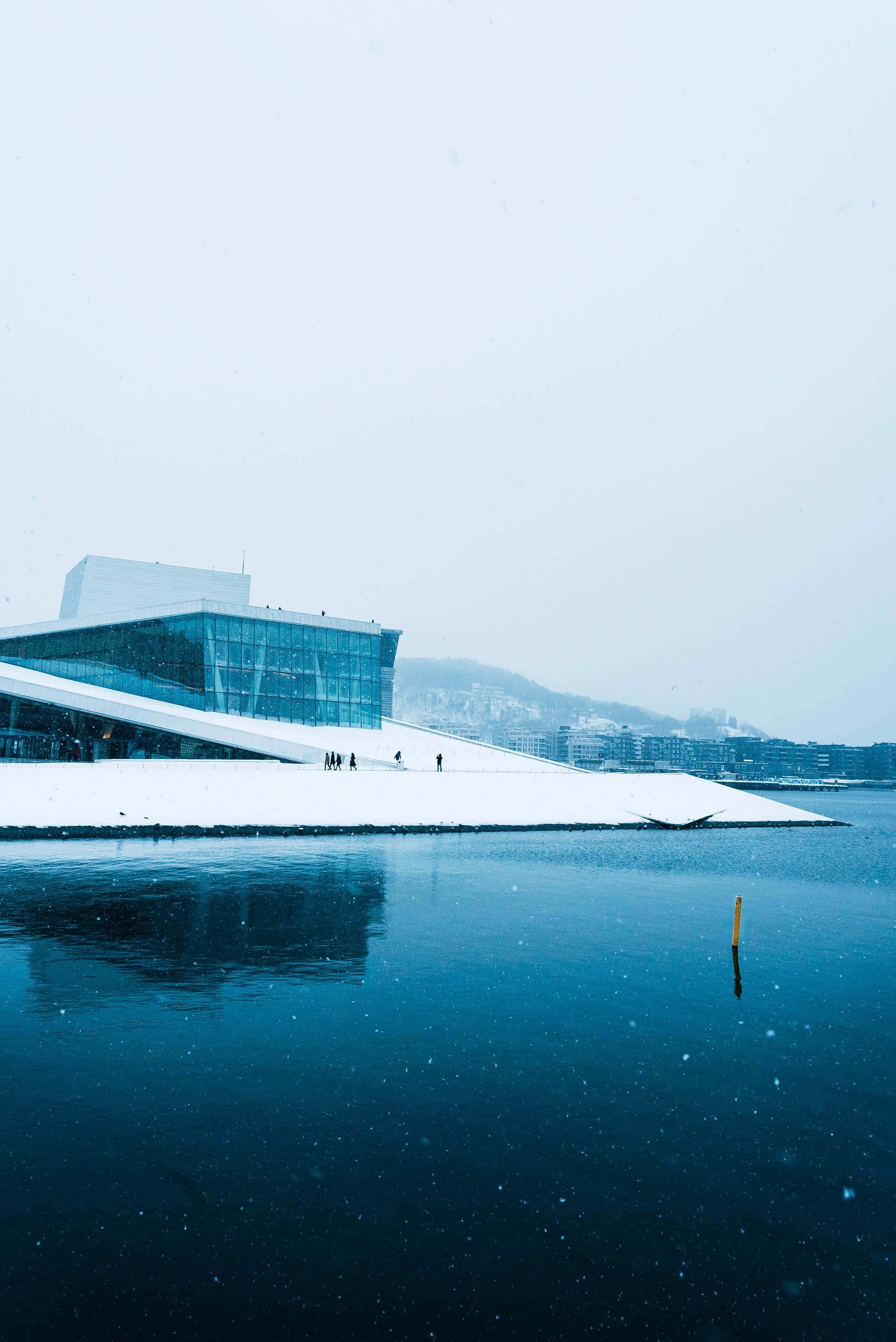 Norway 桌布 壁紙 — 一座現代玻璃和積雪覆蓋的歌劇院在平靜的藍色水面上倒映，背景是白雪皚皚的冬日天空。