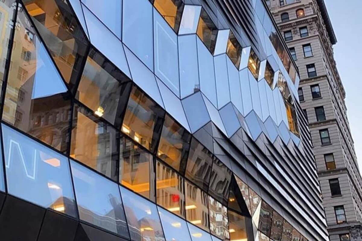 建築 桌布 壁紙 — 一座現代玻璃建築，幾何形切面反射溫暖的室內燈光及相鄰的歷史建築，背景是晴朗的天空。