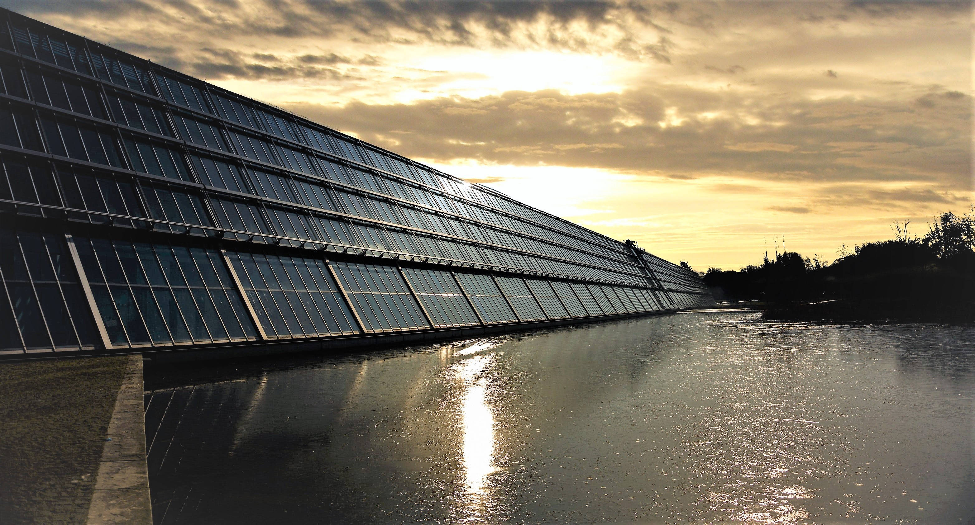 Solar Panel 桌布 壁紙 — 一座現代玻璃建築在平靜的水面上反射出金色夕陽的光芒，天空中有戲劇性的雲彩。