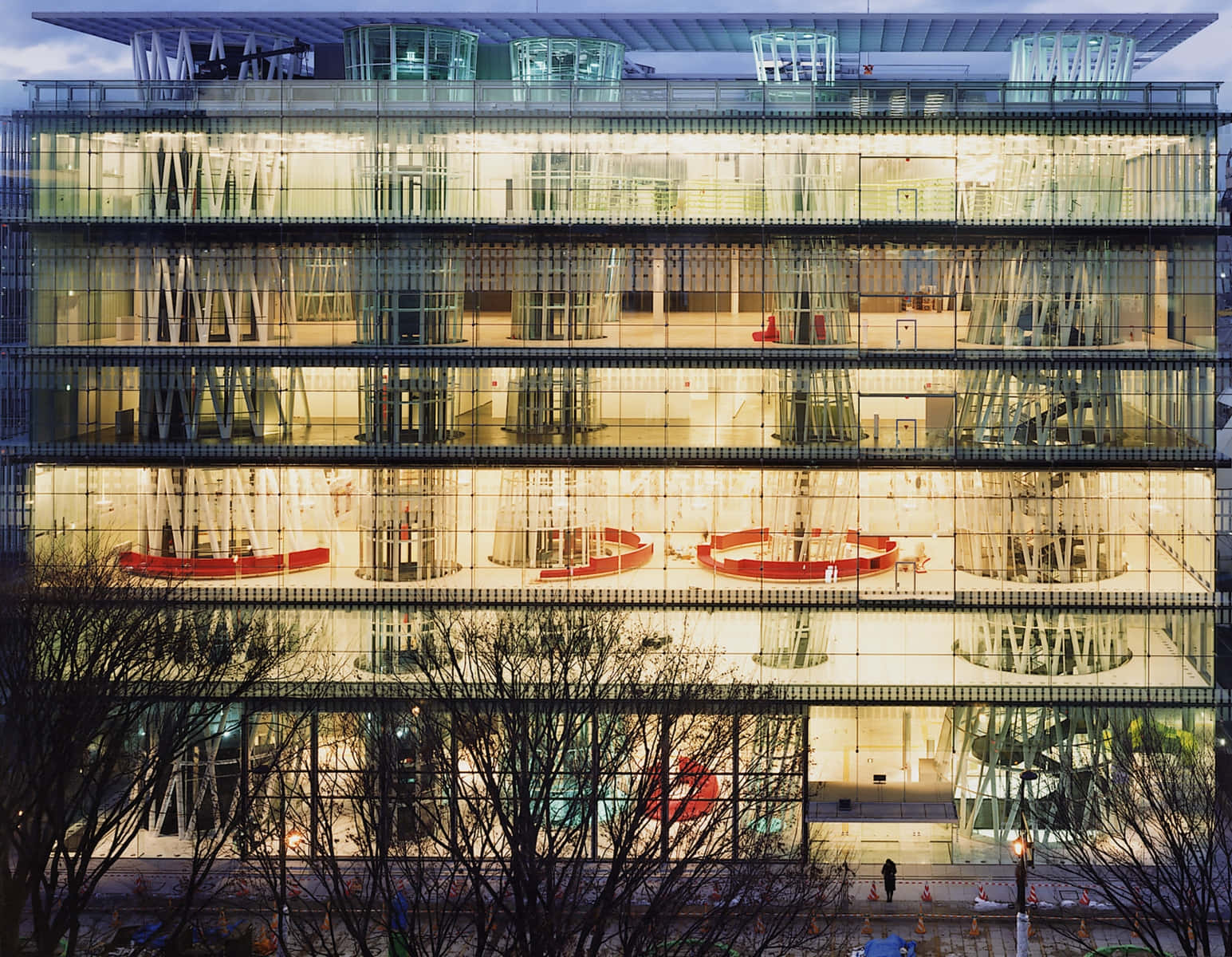 Beautiful Architecture 桌布 壁紙 — 一座現代玻璃建築在黃昏時溫暖地發光，顯露出內部的紅色圓形座位和前景的裸露樹枝。