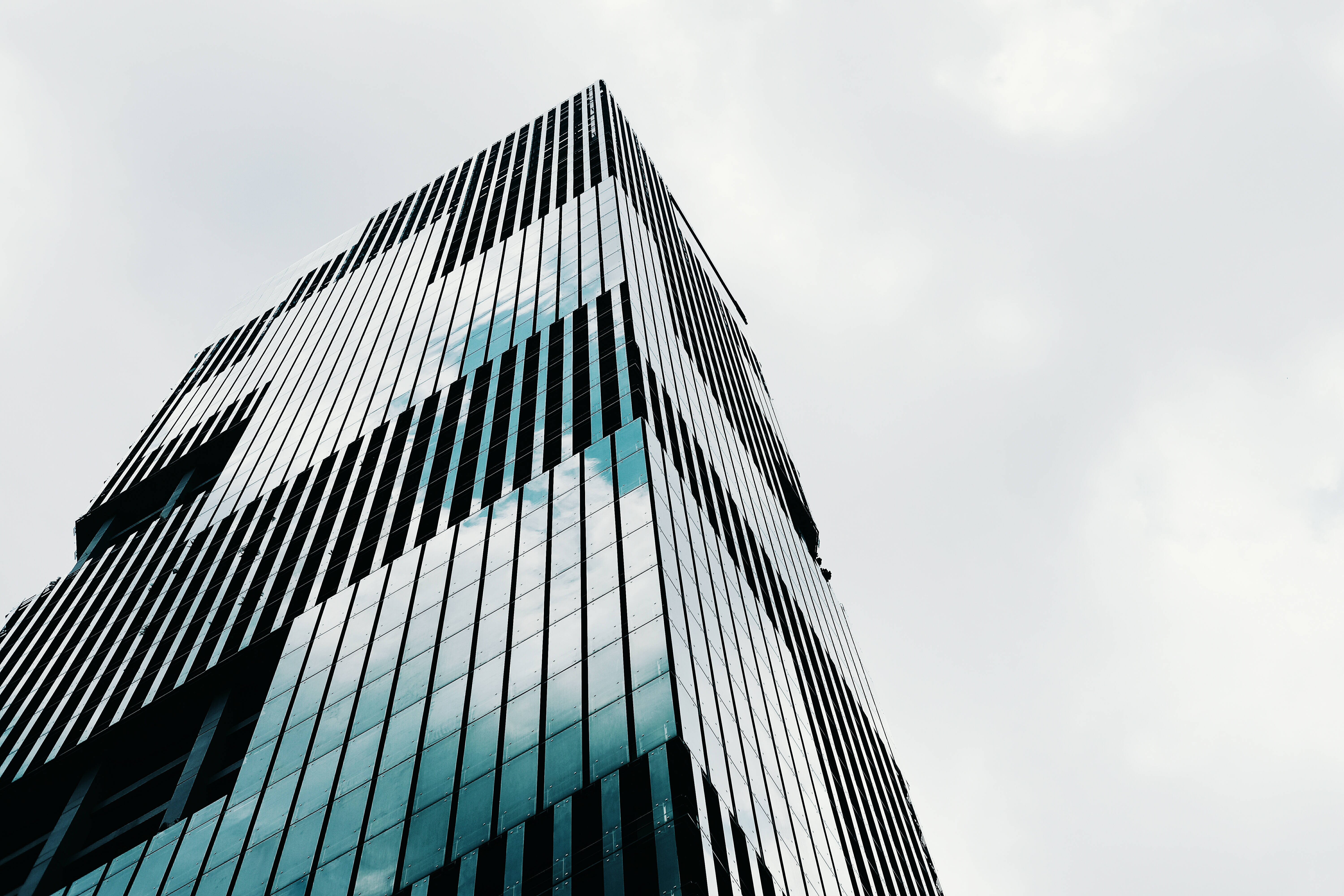 Skyscraper 桌布 壁紙 — 一座現代玻璃摩天大樓，帶有垂直黑色條紋，在淺灰色背景下反射多雲的天空。