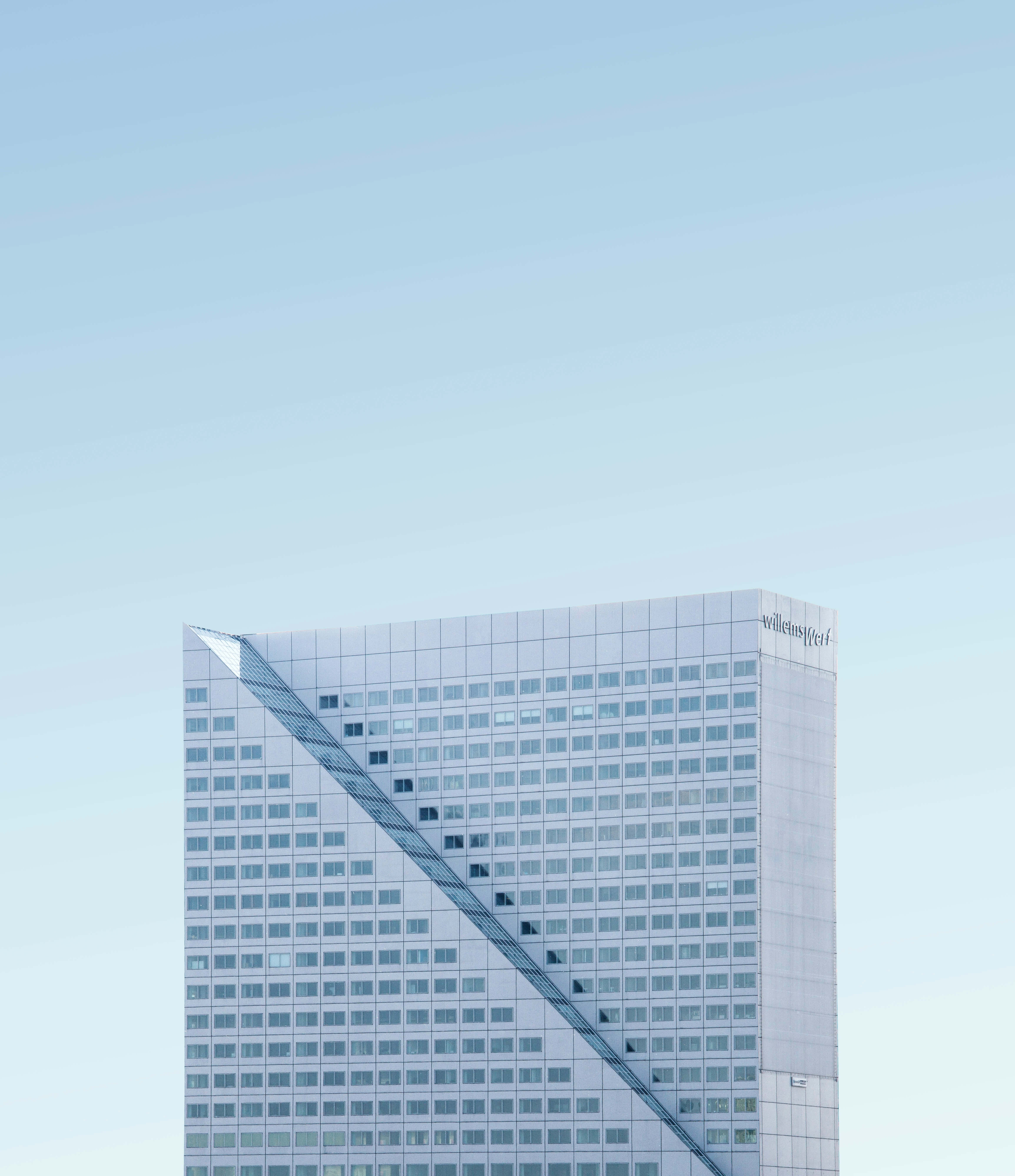 Building 桌布 壁紙 — 一座現代玻璃摩天大樓，帶有對角線玻璃外牆，在清澈的淡藍色天空下聳立。