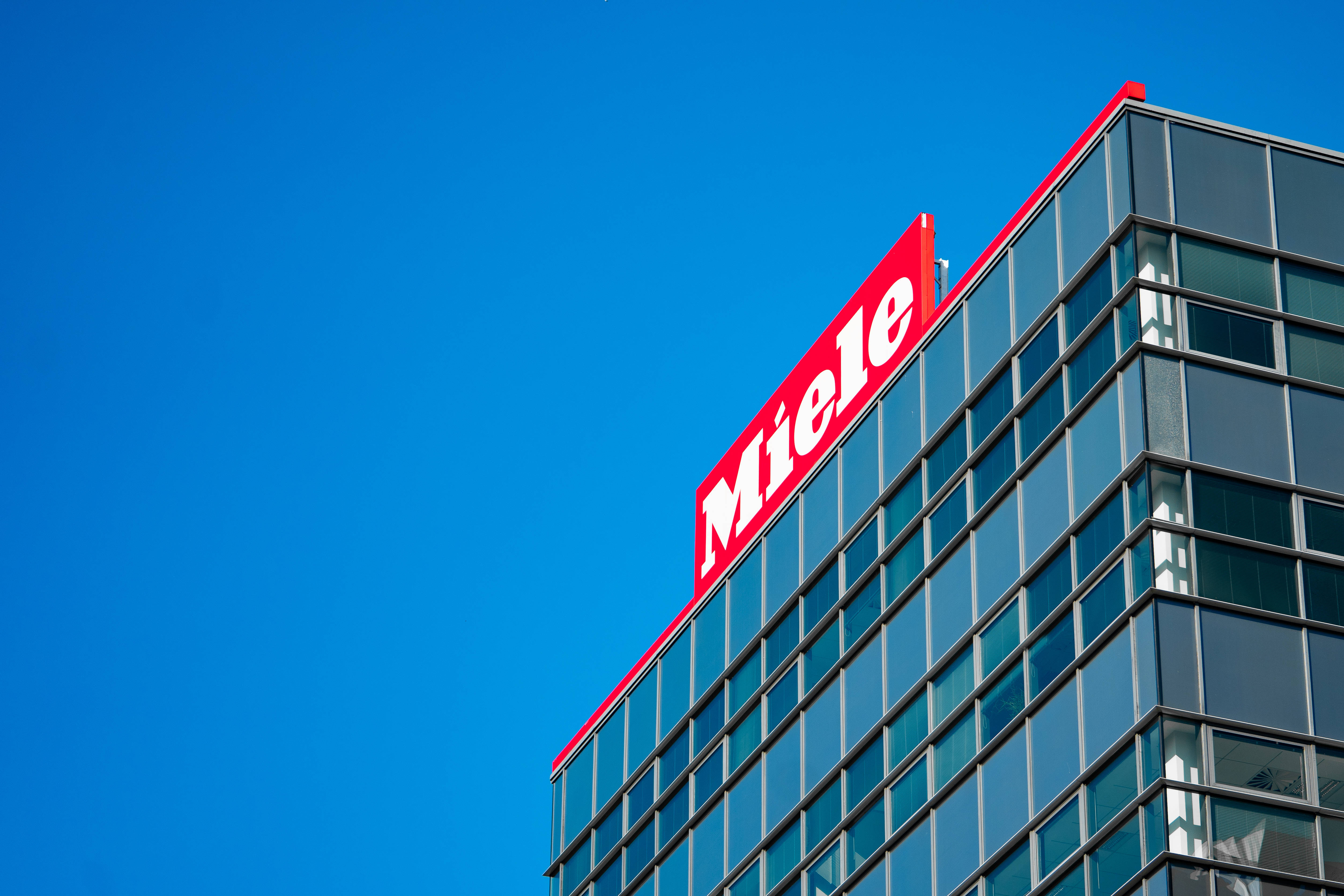 標誌 桌布 壁紙 — 一座現代玻璃摩天大樓，帶有醒目的紅色 Miele 標誌，矗立在清澈的藍天下。