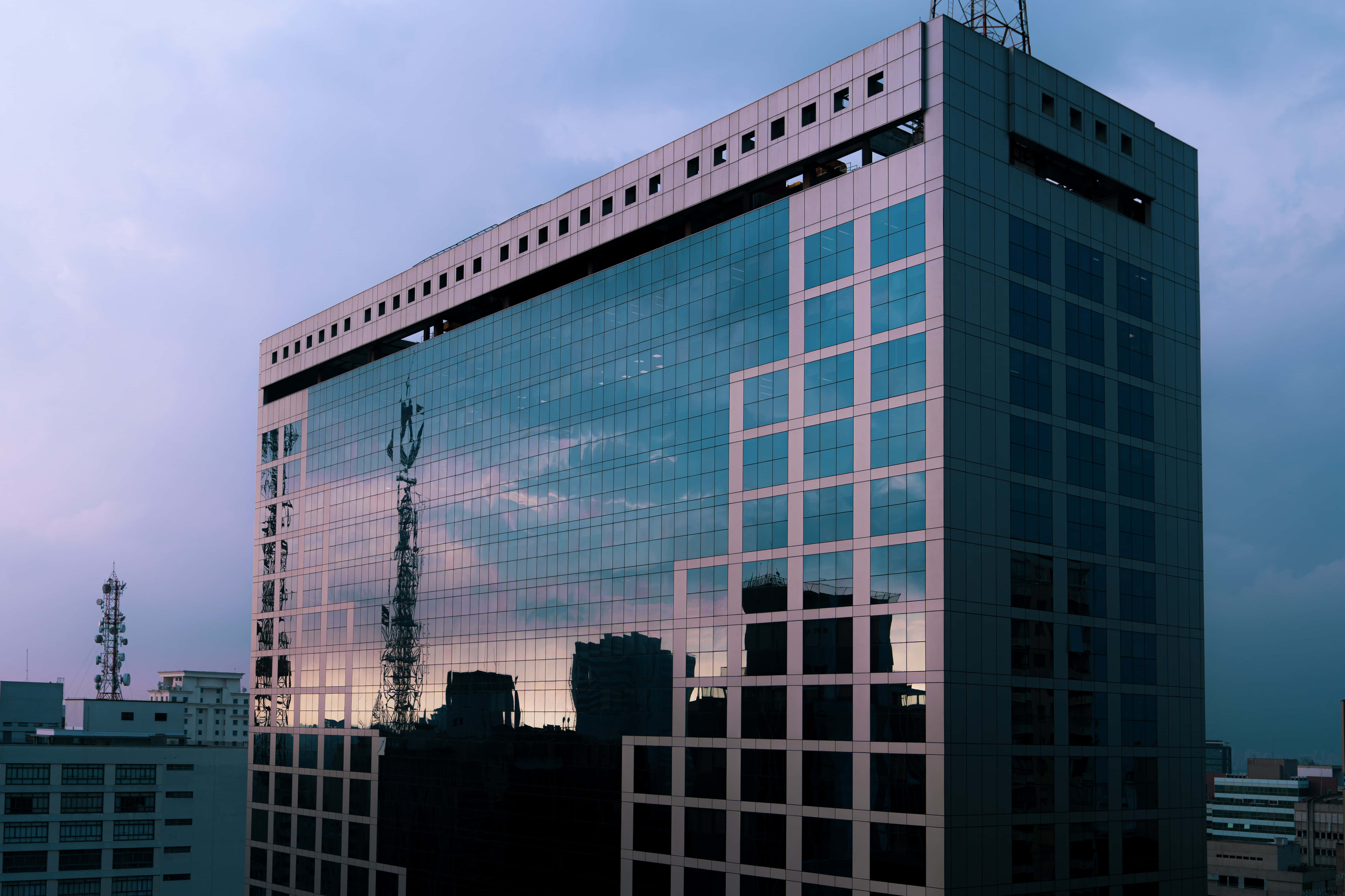 Building 桌布 壁紙 — 一座現代玻璃摩天大樓在其鏡面外牆上反射出暮色天空和周圍的城市景觀。