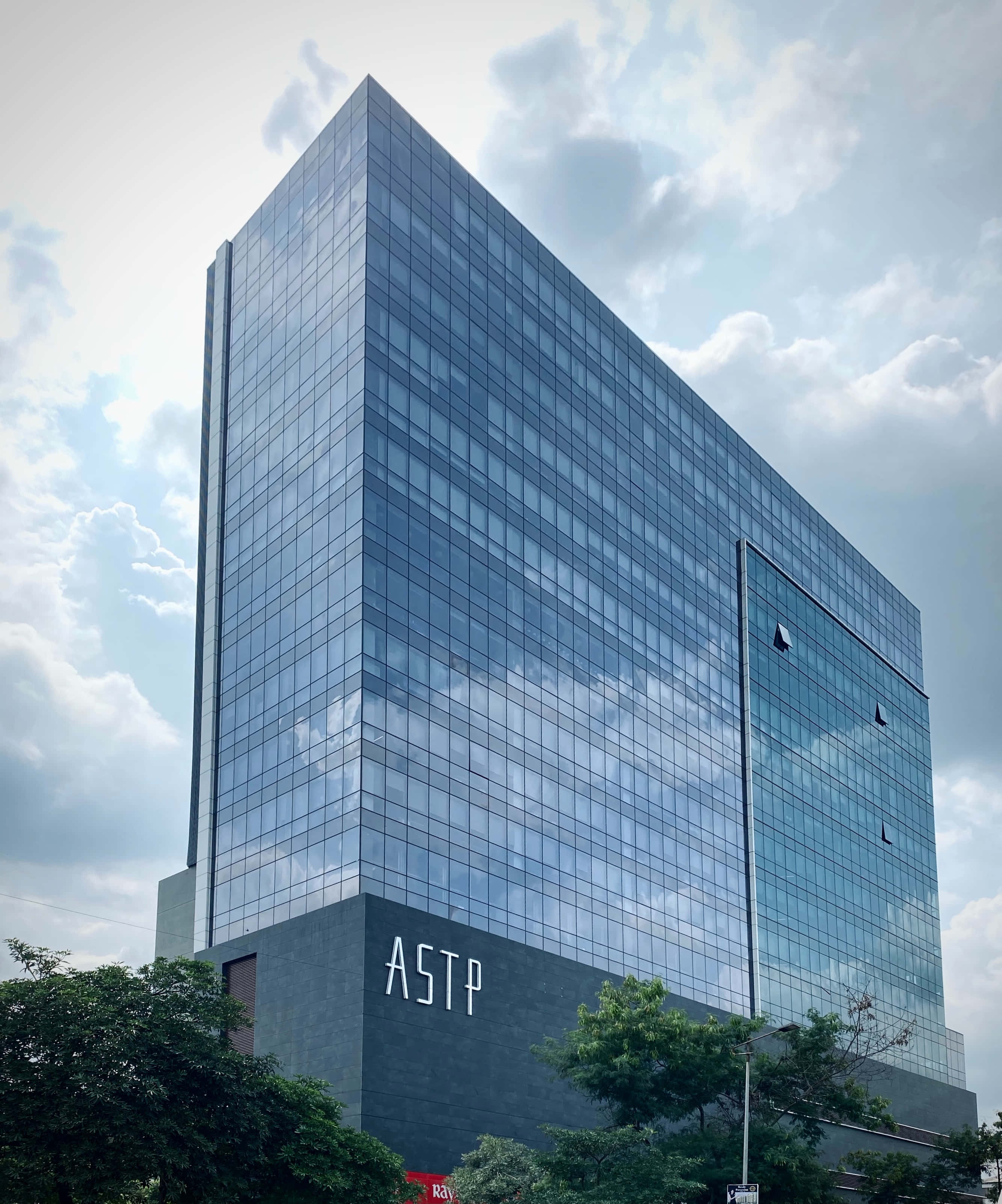 Building 桌布 壁紙 — 一座現代玻璃摩天大樓帶有「ASTP」標誌，在綠色樹木的背景下反射多雲的天空。