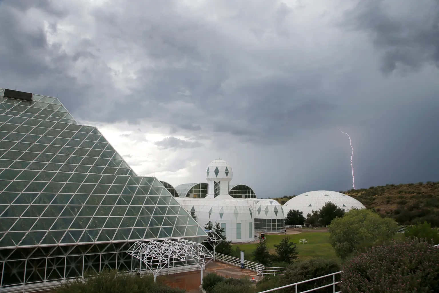 Biosphere 桌布 壁紙 — 一座現代玻璃金字塔和白色圓頂矗立在暴風雨的天空下，遠處有清晰的閃電劃過。