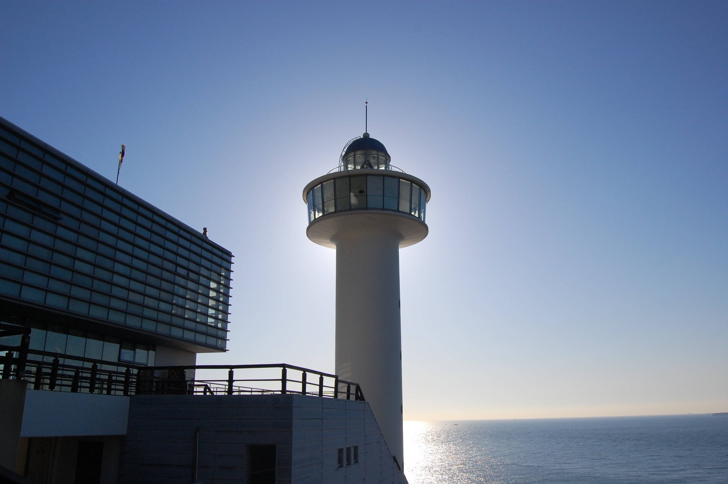 Busan 桌布 壁紙 — 一座現代白色燈塔高聳在玻璃幕牆建築旁，背景是清澈的藍天，夕陽在平靜的海面上落下。
