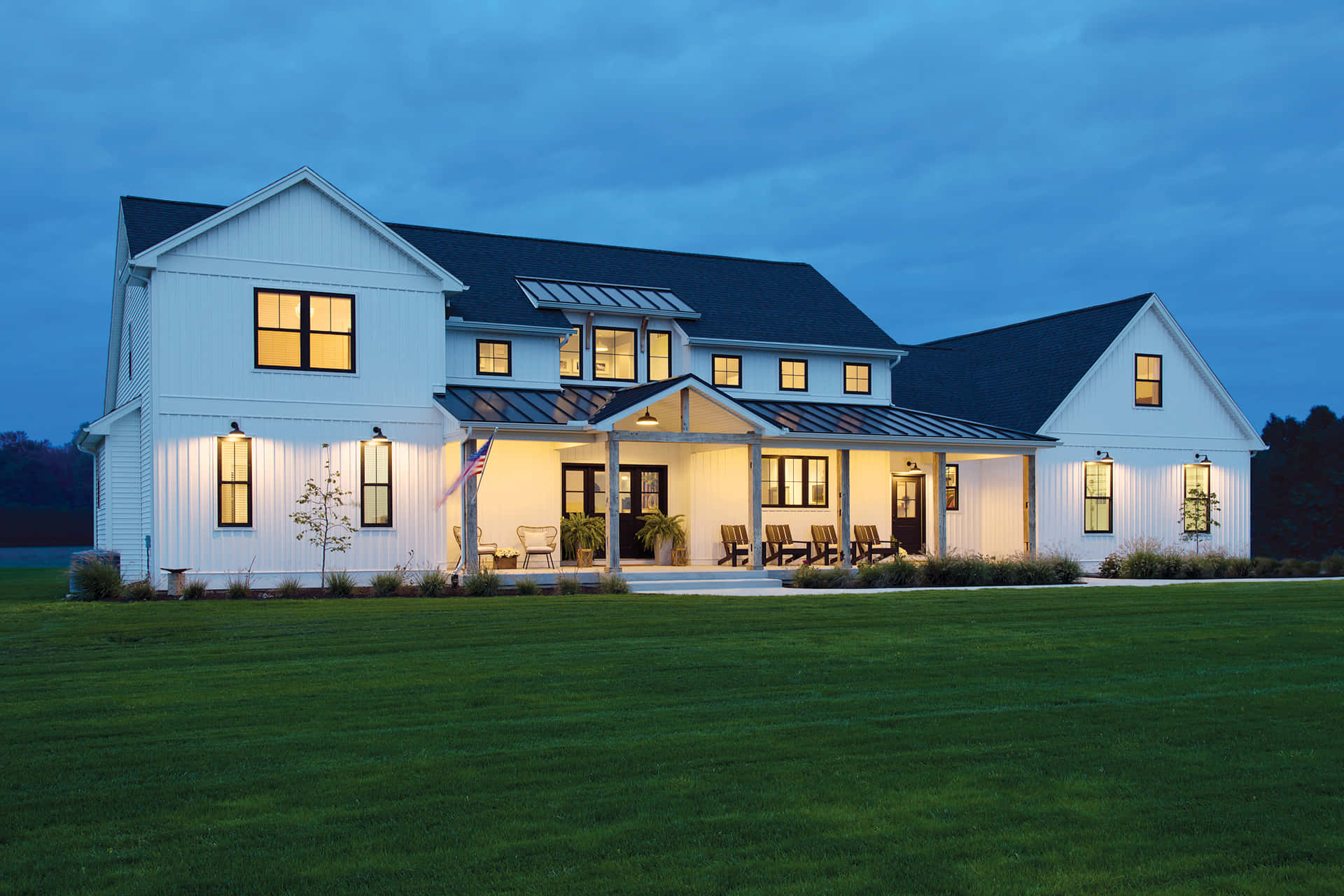 Modern Farmhouse 桌布 壁紙 — 一座現代白色農舍，窗戶發光，在暮色中寧靜地坐落在廣闊的綠色草坪上。