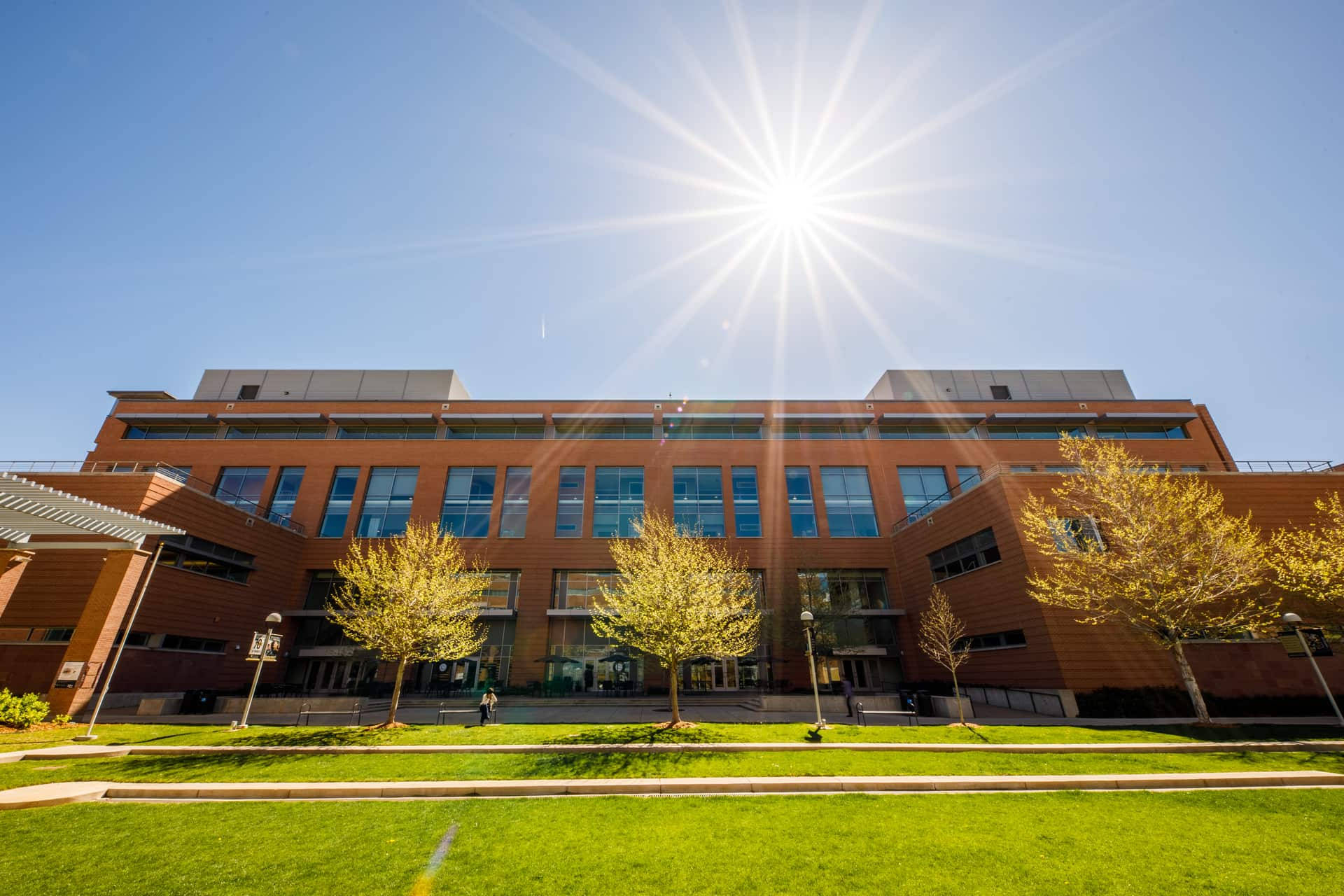 Sunshine 桌布 壁紙 — 一座現代磚砌建築，配有玻璃窗，矗立在明亮的陽光下，周圍環繞著金色的樹木和綠色的草坪。