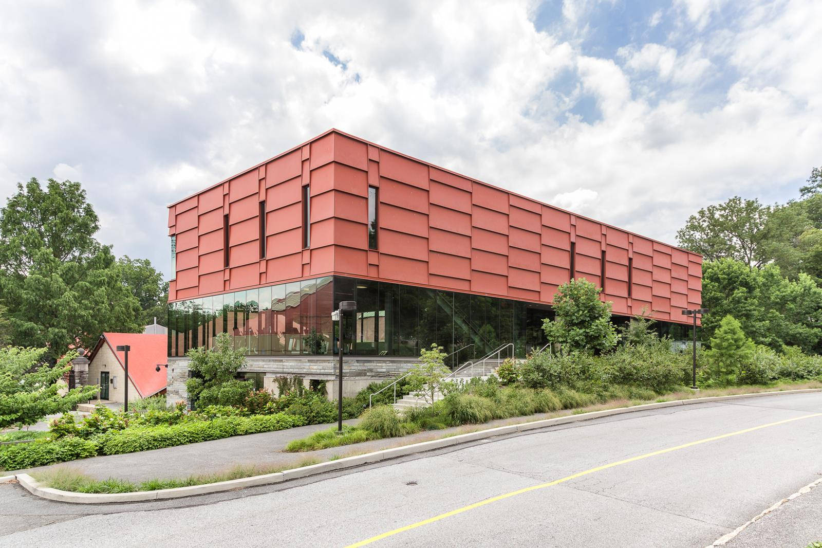 Building 桌布 壁紙 — 一座現代紅色幾何建築，玻璃幕牆，位於路邊，天空多雲，周圍環繞著綠色樹木和灌木。