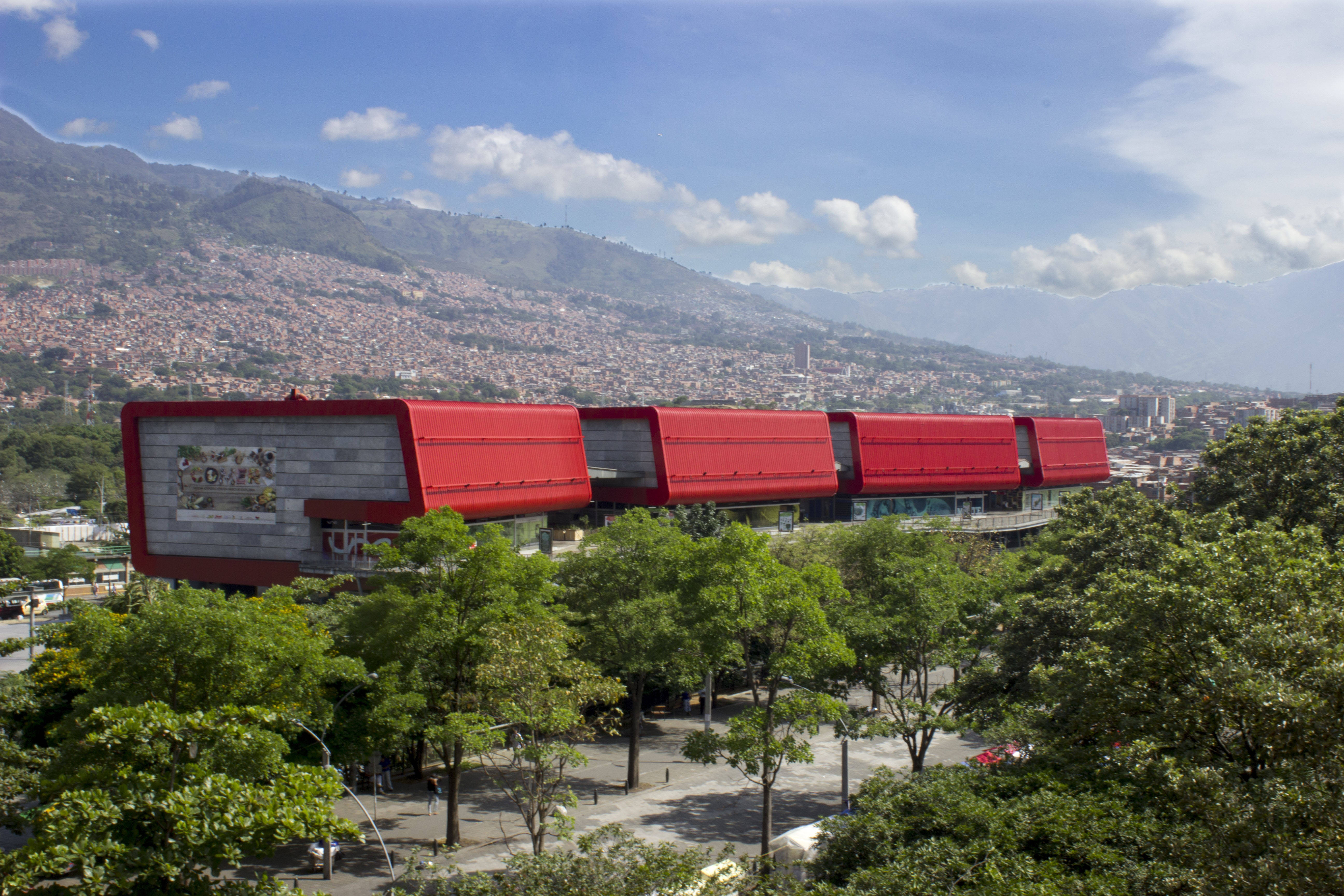 Medellin 桌布 壁紙 — 一座現代紅頂建築，隱藏在綠樹叢中，背後是廣闊的山城景觀，天空湛藍。