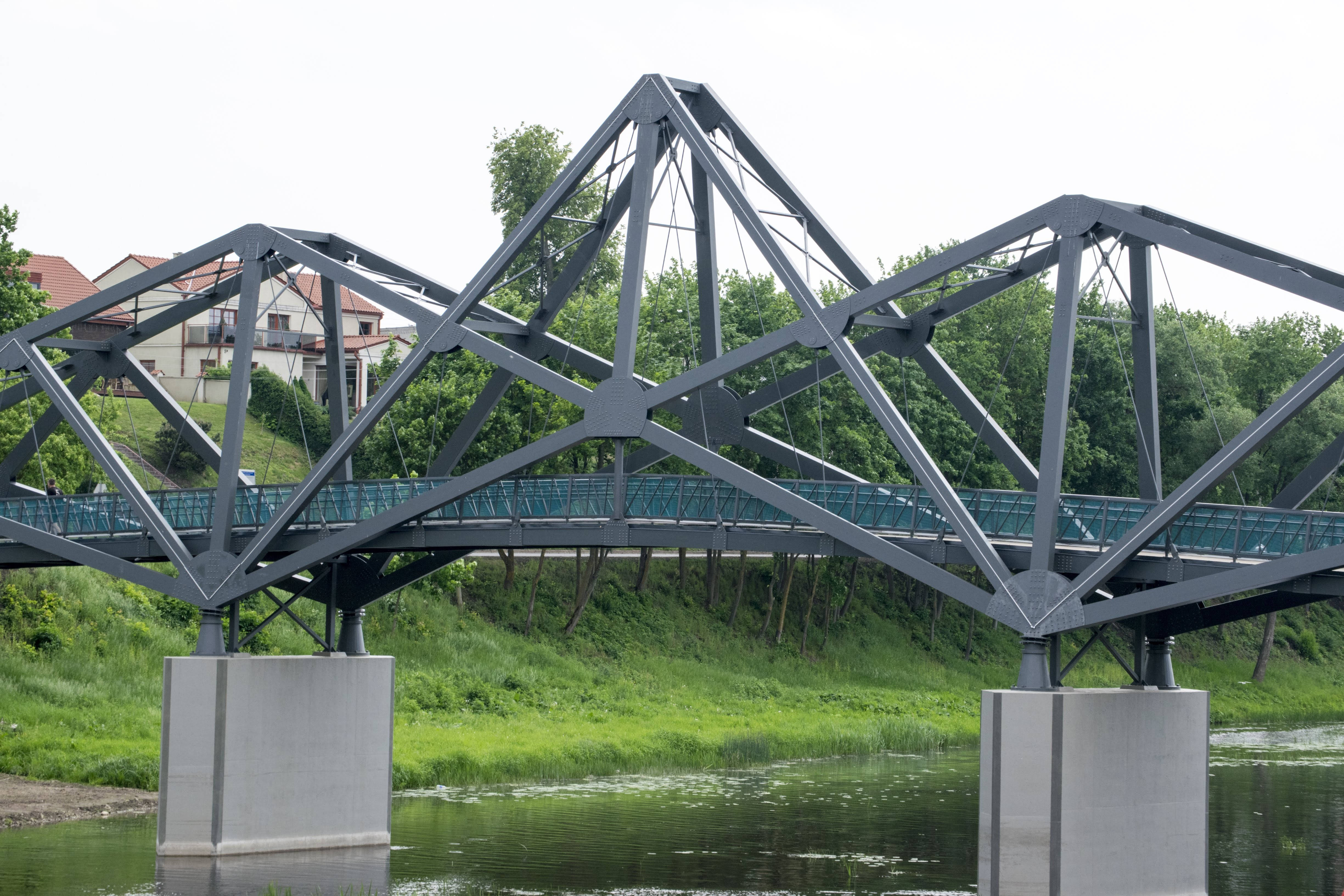Lithuania 桌布 壁紙 — 一座現代鋼桁架橋橫跨一條平靜的河流，兩岸綠樹成蔭，背景可見房屋。