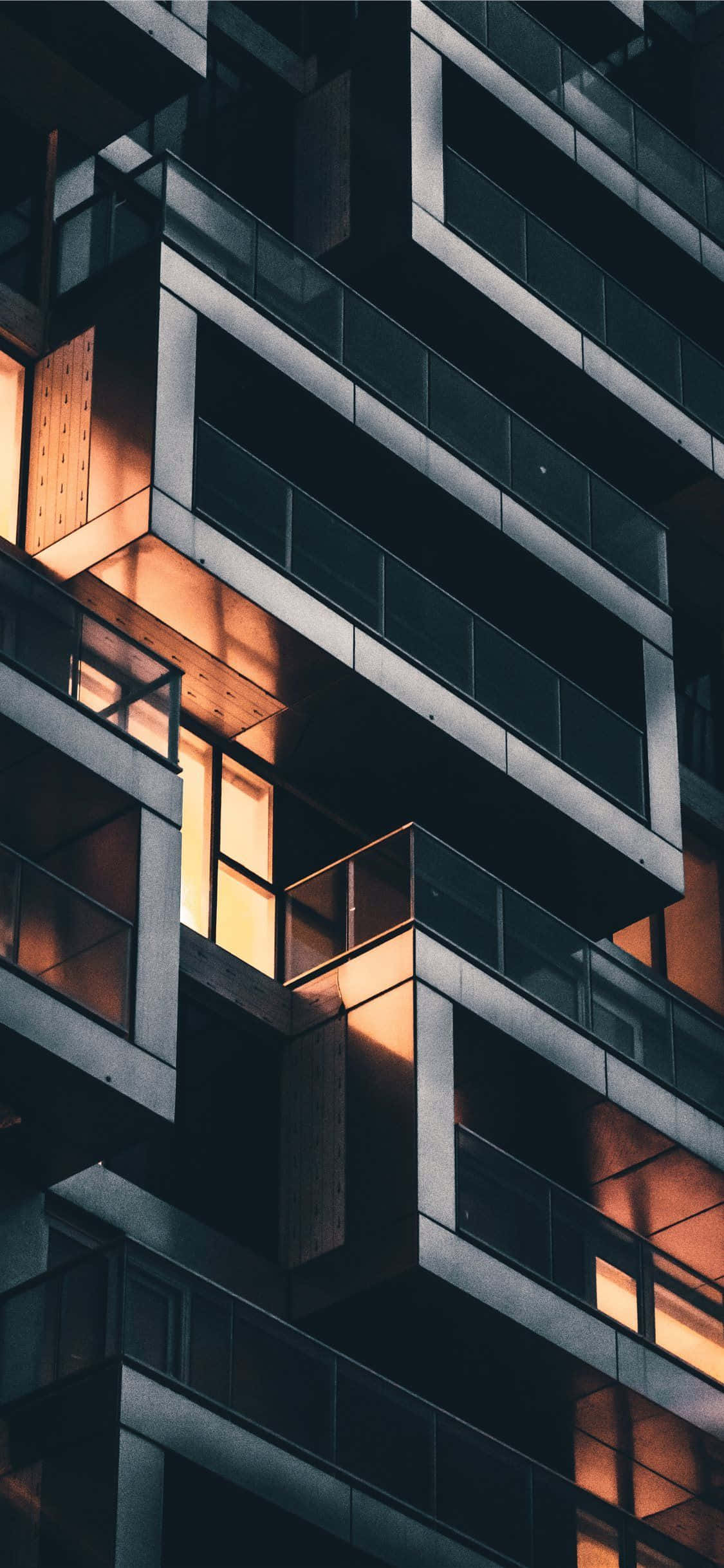 建築 桌布 壁紙 — 一座現代高層建築在黃昏時分，溫暖的室內燈光透過幾何窗戶照亮黑暗的外牆。