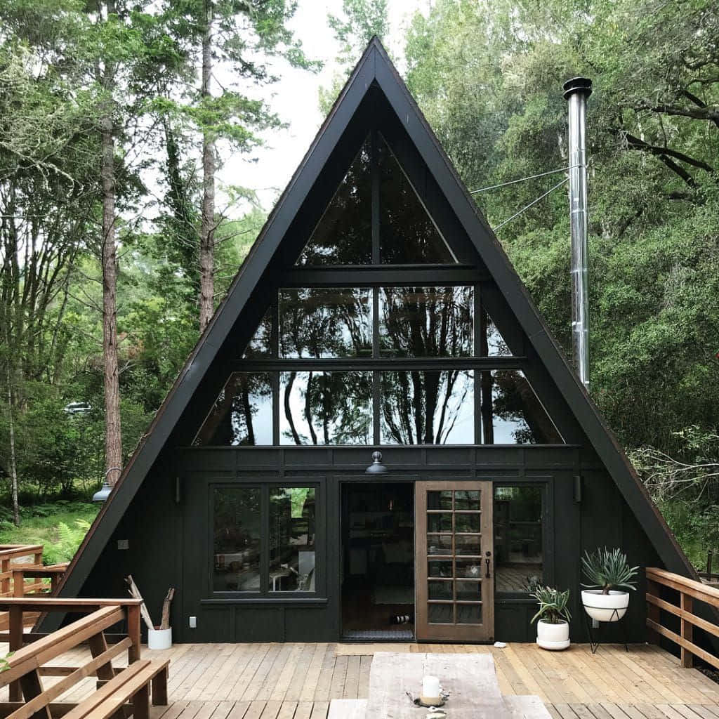 Cottage 桌布 壁紙 — 一座現代黑色A字形小屋，帶有大玻璃窗，坐落在高聳綠樹間的木製平台上。