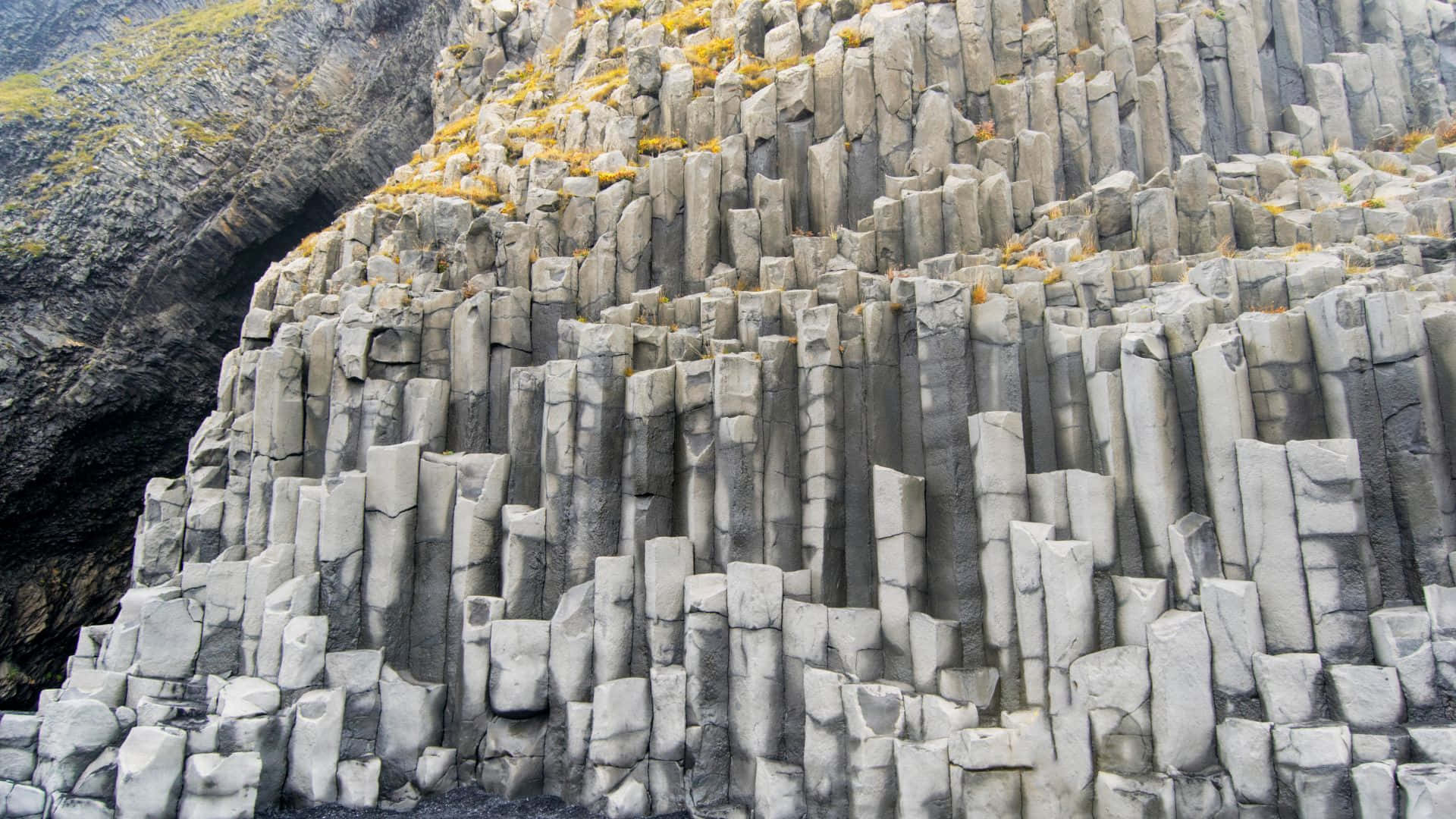冰島 桌布 壁紙 — 一座由六角形玄武岩柱組成的 towering 懸崖從深色火山岩中陡峭升起，裂縫中附著稀疏的黃綠色苔蘚。