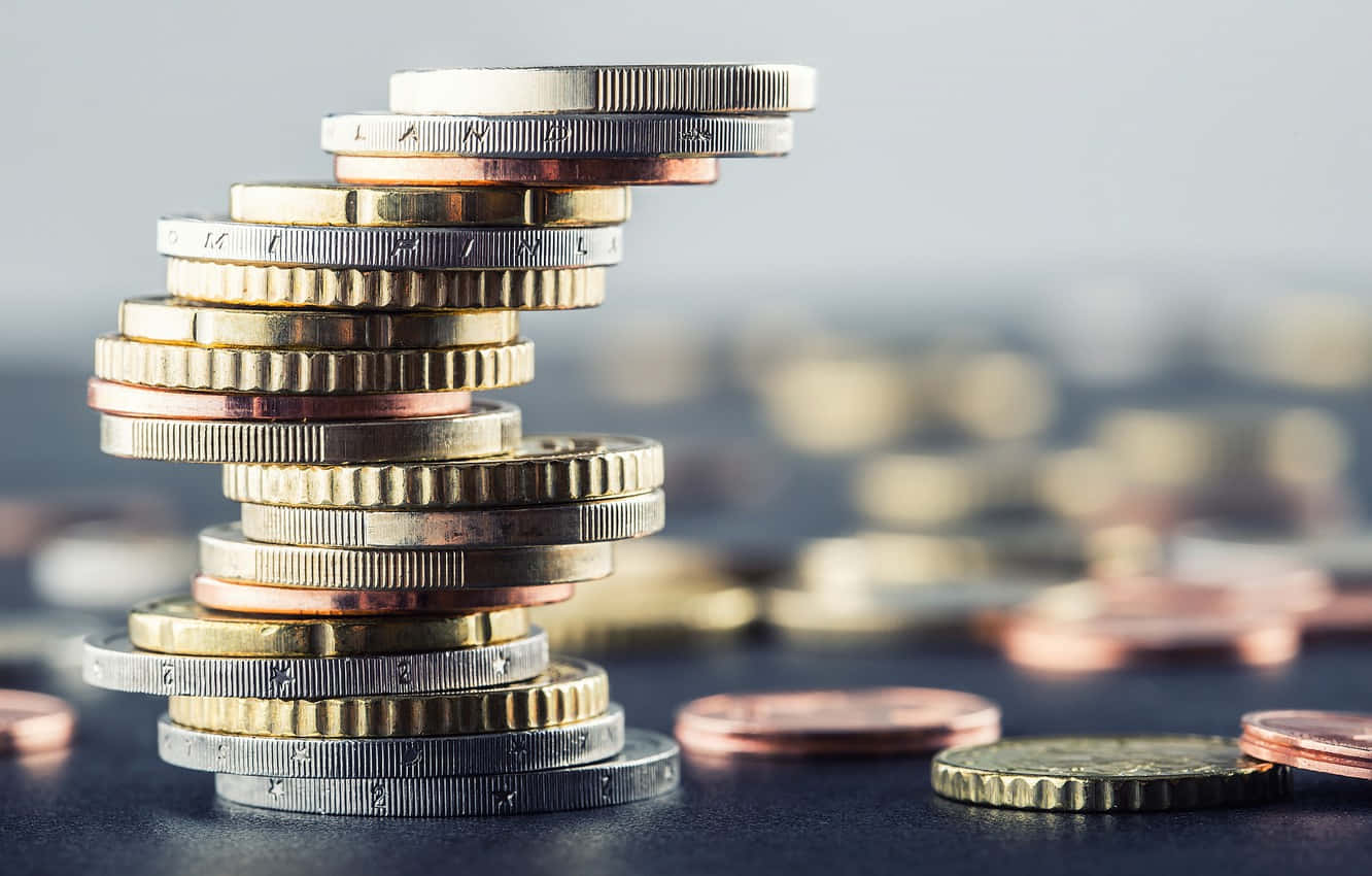 金錢 桌布 壁紙 — 一座由歐洲硬幣堆疊而成、搖搖欲墜的螺旋塔，在模糊的散落貨幣背景襯托下聳立。