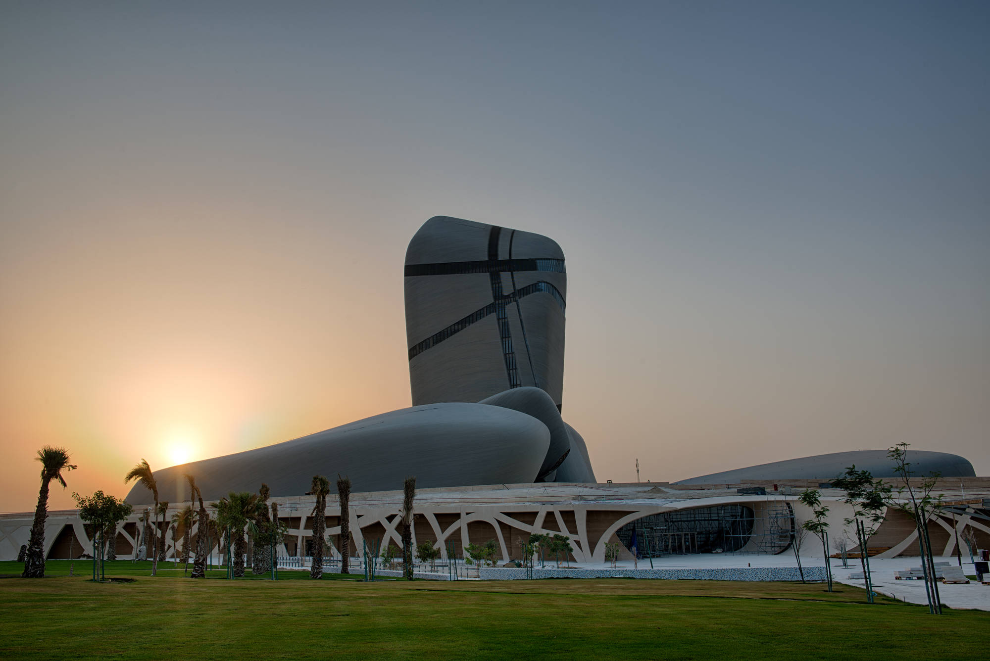 Saudi 桌布 壁紙 — 一座由玻璃和鋼材建造的未來主義曲線建築，在溫暖的日落天空下矗立，前景有棕櫚樹。