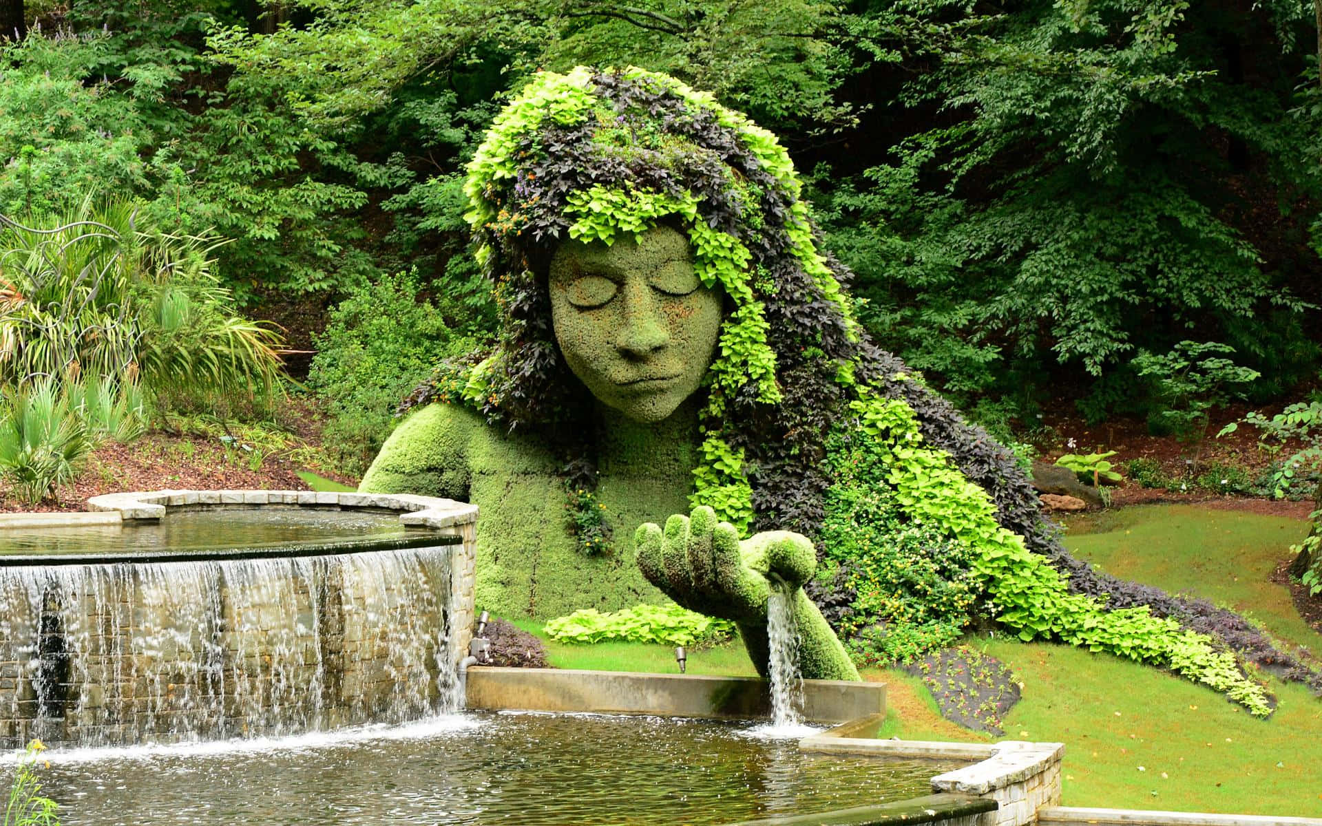 Botanical Garden 桌布 壁紙 — 一座由綠色樹葉製成的女性花園雕塑，寧靜地將水從手中輕輕倒入一個被茂密樹木環繞的石頭噴泉中。