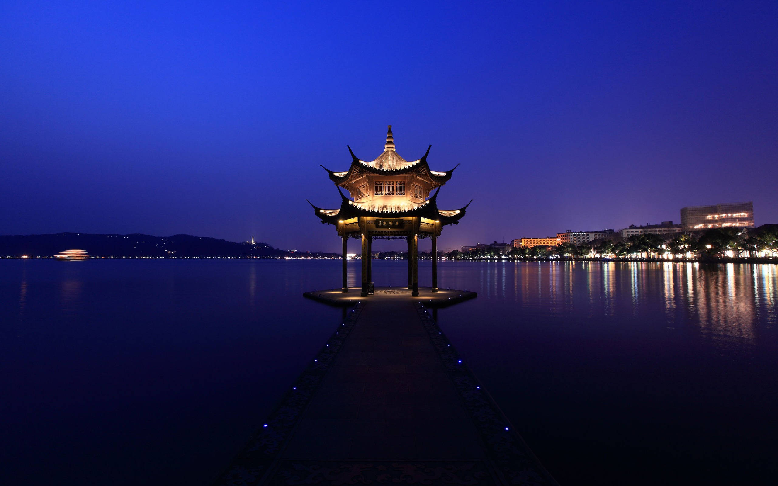 Hangzhou 桌布 壁紙 — 一座發光的傳統中式亭子矗立在寧靜湖面上的碼頭盡頭，在深藍色的暮色天空下。