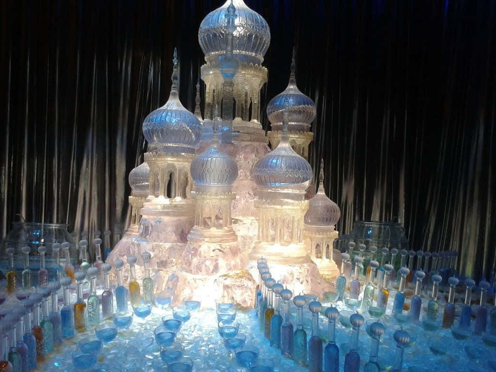 Ice Sculpture 桌布 壁紙 — 一座發光的冰城堡，帶有精緻的圓頂和尖塔，周圍環繞著藍色玻璃瓶，位於反光表面上。