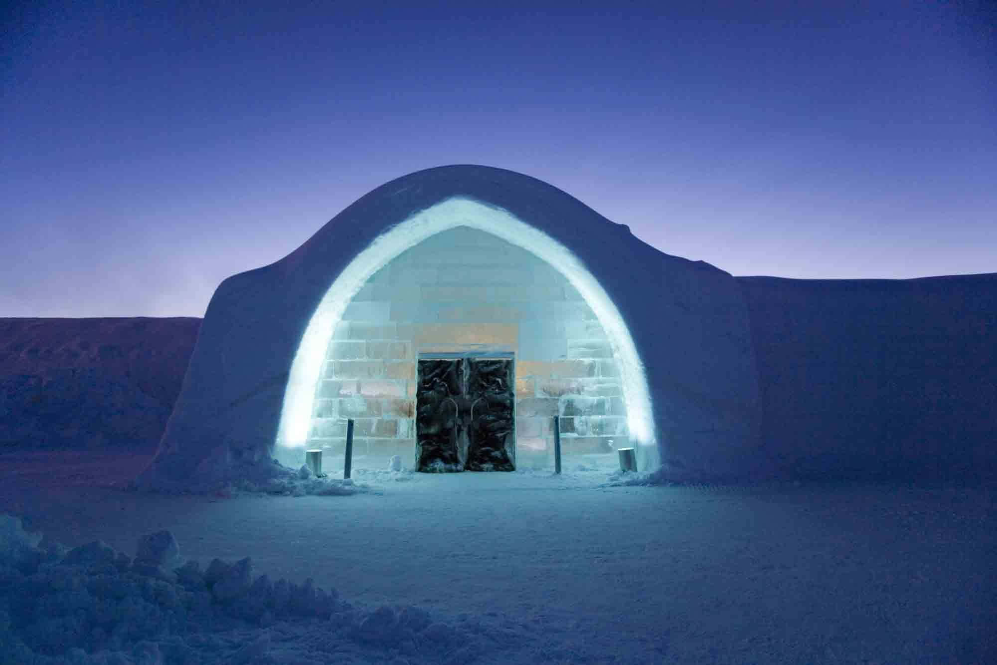 Ice Hotel 桌布 壁紙 — 一座發光的冰屋，帶有拱形入口和雕刻的門，矗立在深藍色天空下的雪黃昏中。