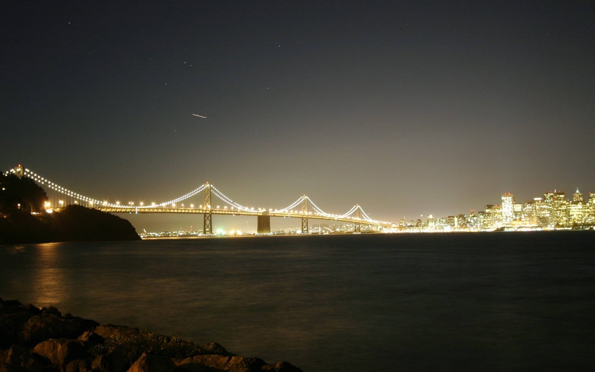 View 桌布 壁紙 — 一座發光的吊橋橫跨黑暗的海灣，夜空繁星點點，岸邊的城市燈光閃爍。