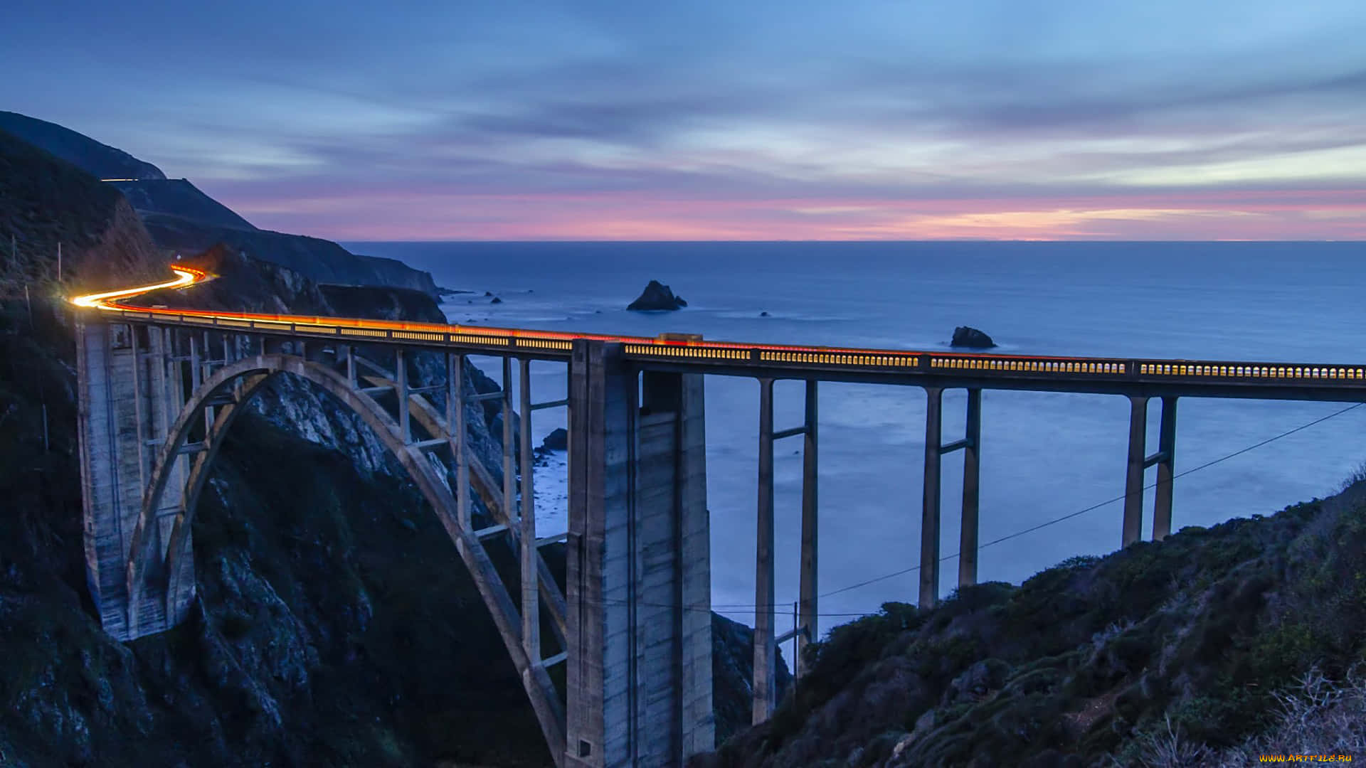 macOS Big Sur 動態桌布 桌布 壁紙 — 一座發光的橋樑在暮色中拱起於平靜的海洋之上，汽車燈光的條紋在紫色和粉紅色的天空襯托下勾勒出它的曲線。