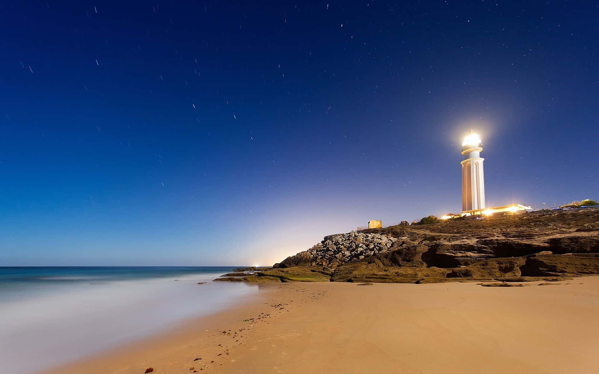 Seaside 桌布 壁紙 — 一座發光的燈塔矗立在岩石懸崖上，下方是星空夜空和模糊平滑的海洋。