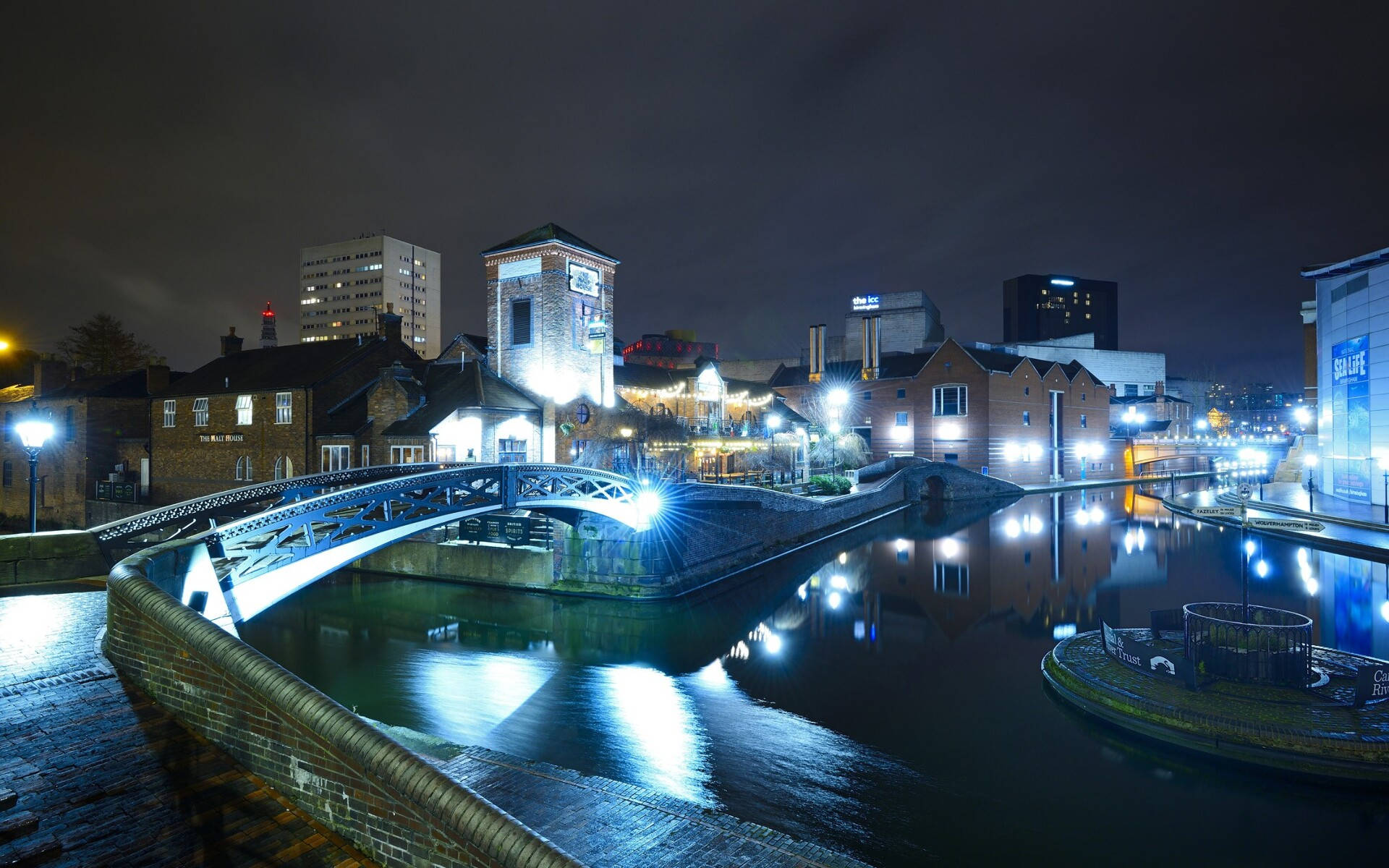 Birmingham 桌布 壁紙 — 一座發光的現代橋樑在夜晚拱跨平靜的運河，在黑暗的天空下映照著城市燈光和磚砌建築。