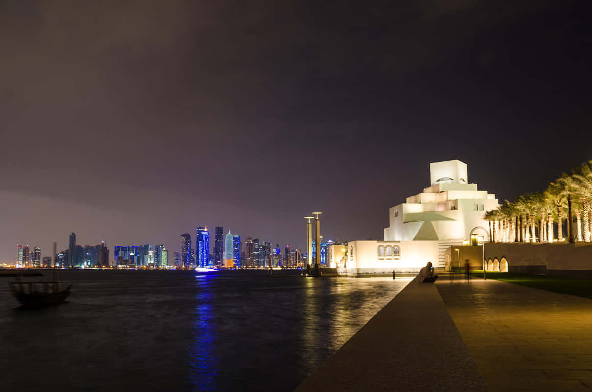 Museum Of Islamic Art 桌布 壁紙 — 一座發光的白色博物館，採用幾何建築，夜晚矗立於水邊，在黑暗的天空下反射城市燈光和棕櫚樹。