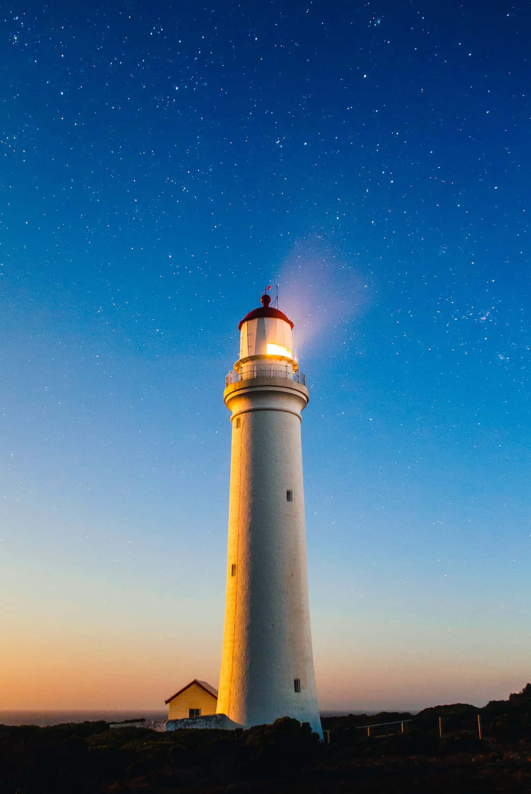 Lighthouse 桌布 壁紙 — 一座發光的白色燈塔高聳在星空暮色中，地平線泛著橙色光芒。