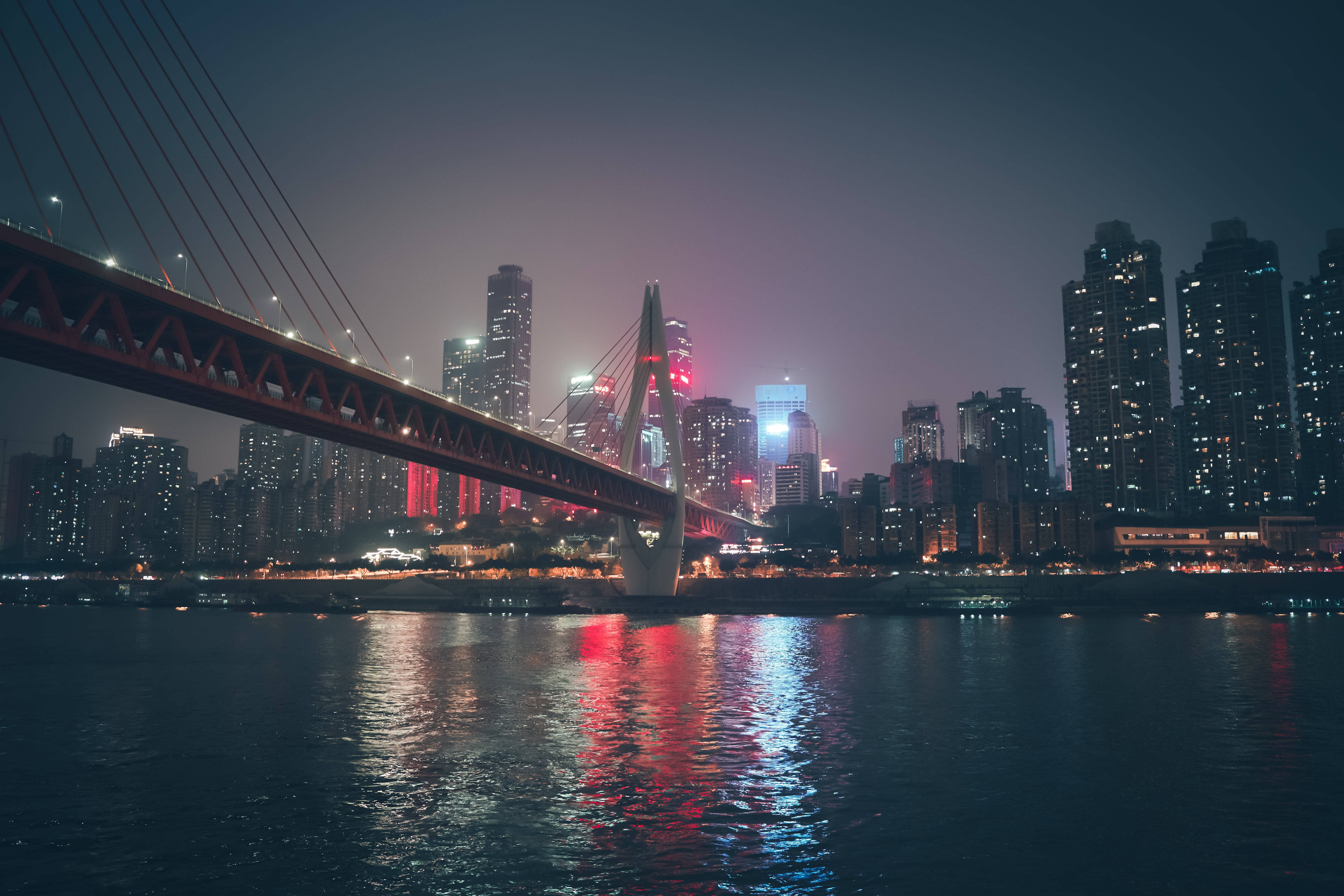 Chongqing 桌布 壁紙 — 一座發光的紅色吊橋橫跨黑暗的河流，映照著霓虹城市燈光，背景是朦朧的夜空。