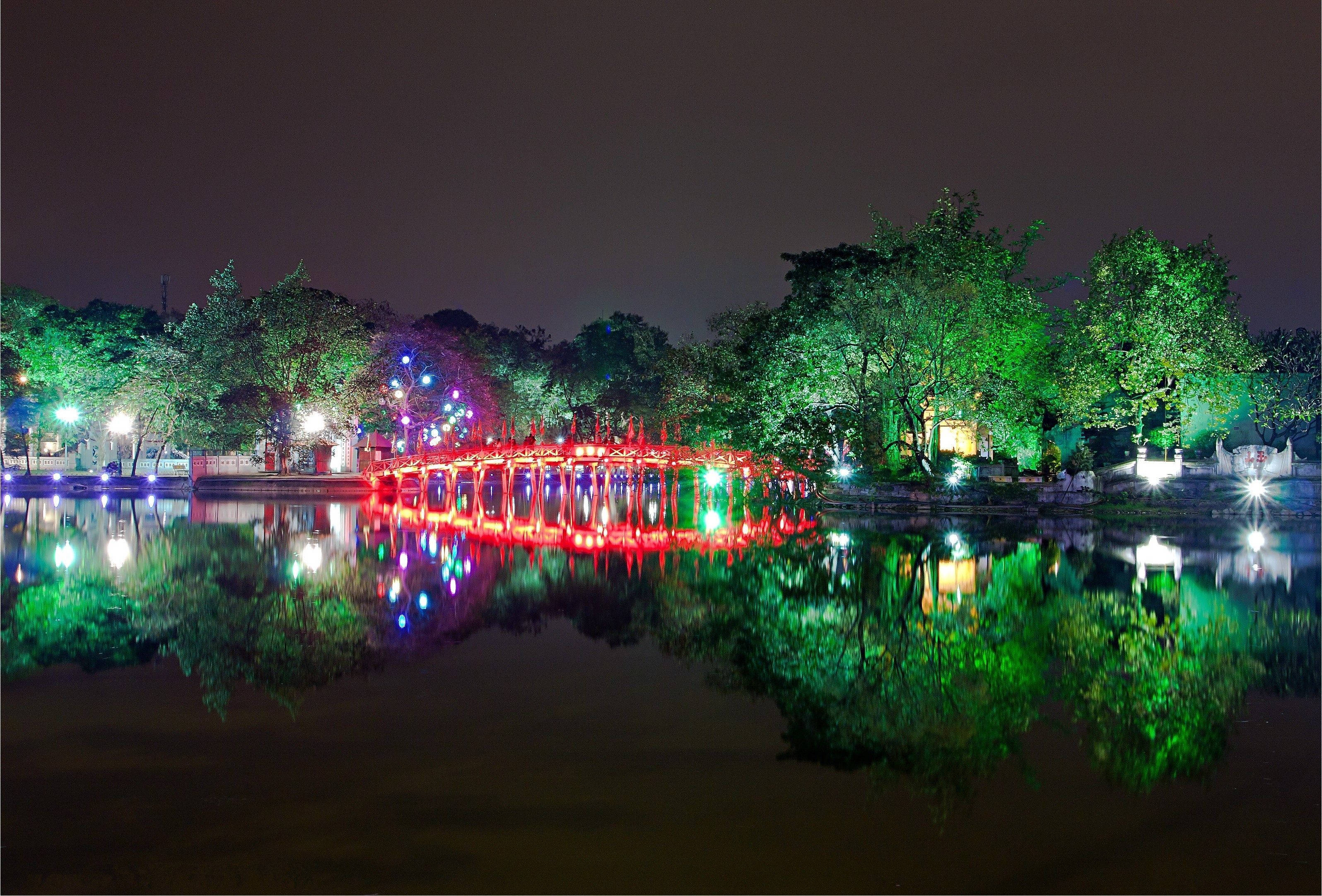 Hanoi 桌布 壁紙 — 一座發光的紅色拱橋在平靜的水面上，映照出五彩燈光和綠樹的充滿活力的夜景。