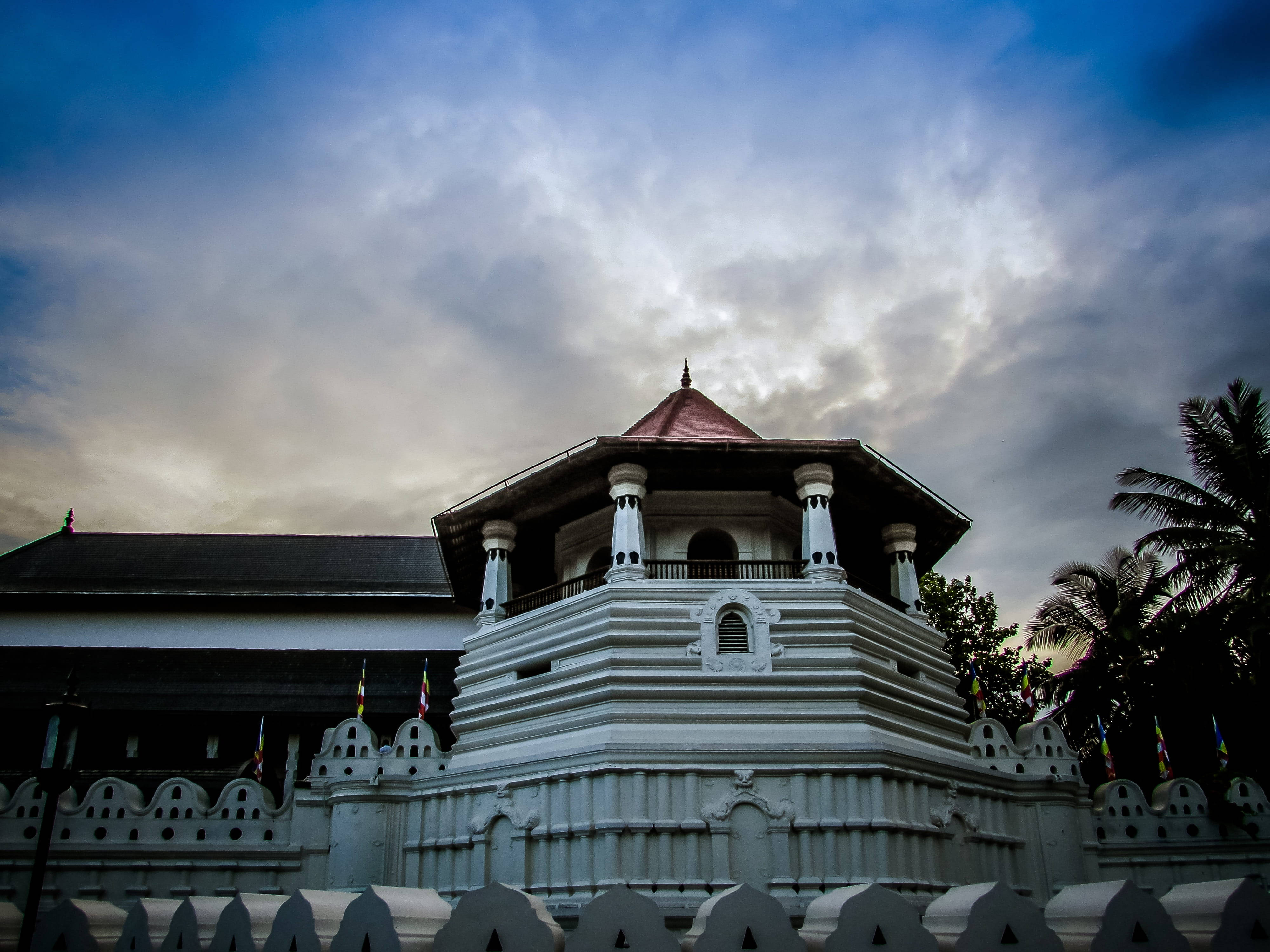 Sri Lanka 桌布 壁紙 — 一座白色分層寺廟，紅色屋頂，矗立在戲劇性的多雲天空下，附近有棕櫚樹。