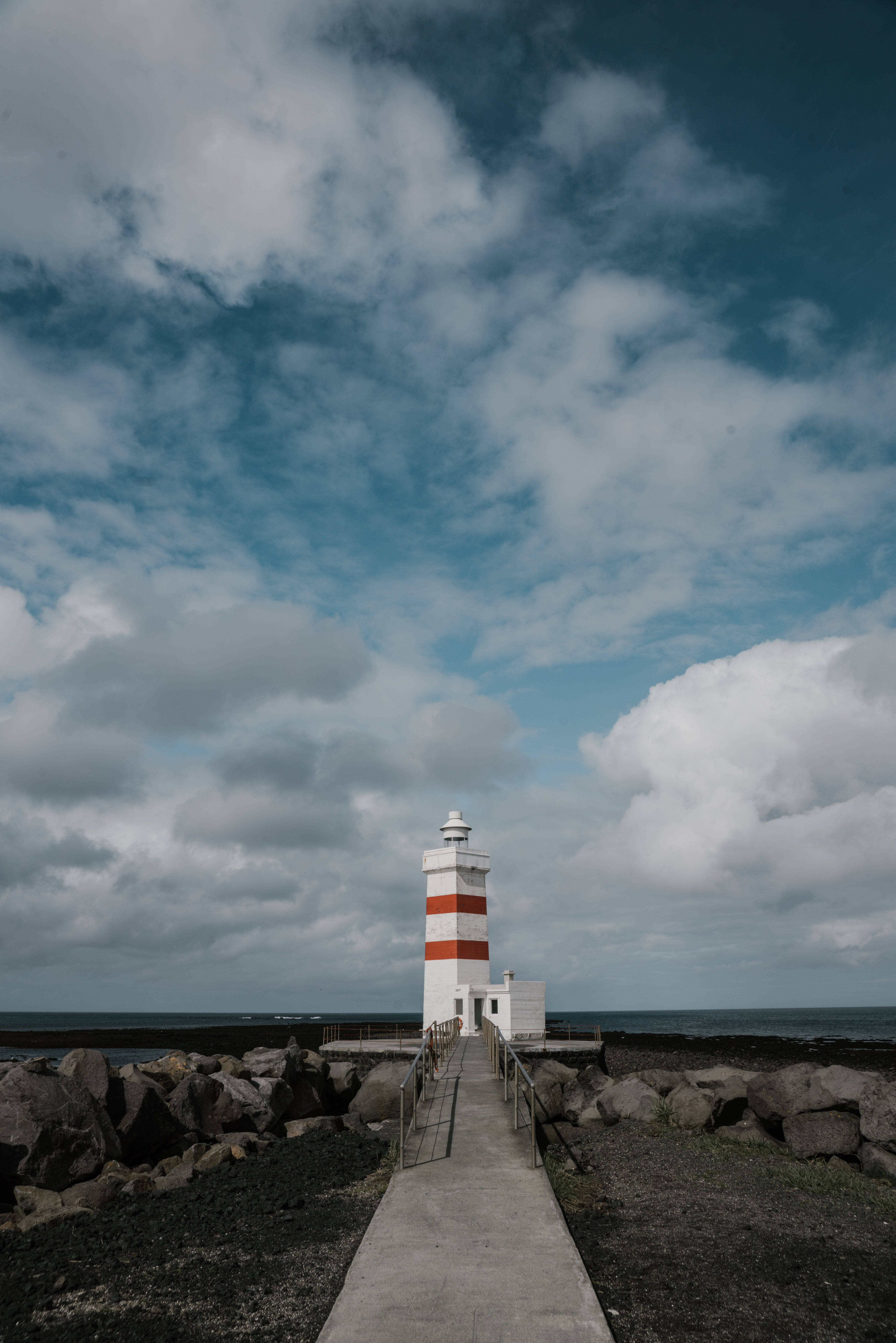Clouds Aesthetics 桌布 壁紙 — 一座白色和紅色條紋的燈塔矗立在混凝土小徑的盡頭，周圍是深色火山岩，上方是戲劇性的多雲天空。