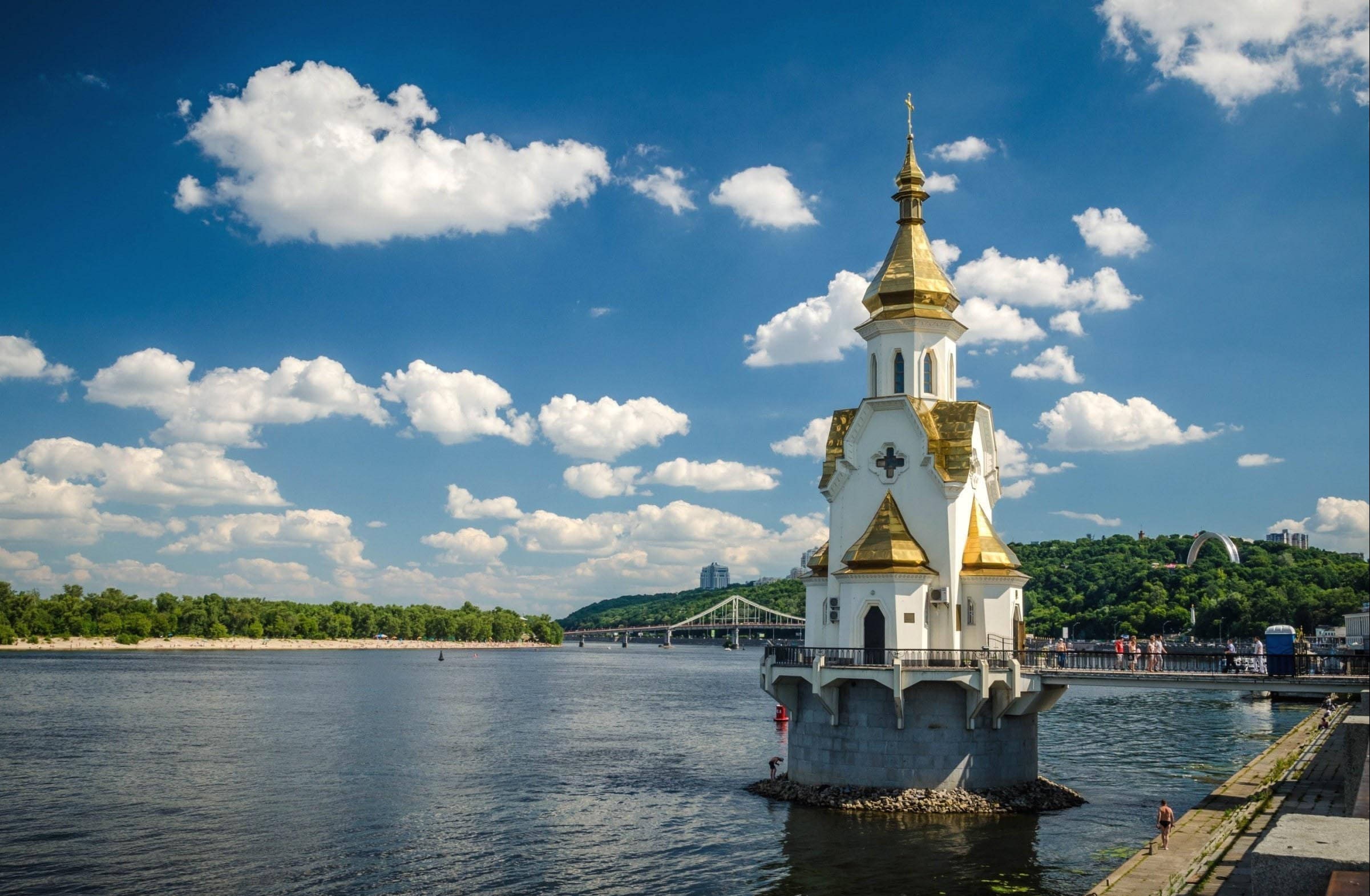 Ukraine 桌布 壁紙 — 一座白色和金色的東正教教堂矗立在寬闊河流中央的石基上，上方是晴朗的藍天和蓬鬆的白雲。