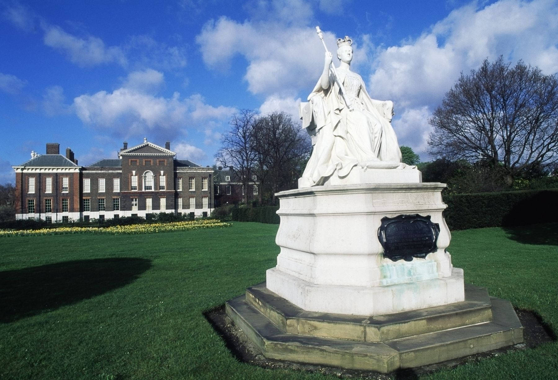 England 桌布 壁紙 — 一座白色大理石雕像，一位戴著王冠的女王手持權杖，立於基座上，位於修剪整齊的綠色草坪上，背景是一座宏偉的磚砌豪宅和黃色鬱金香，在明亮的藍天白雲下。